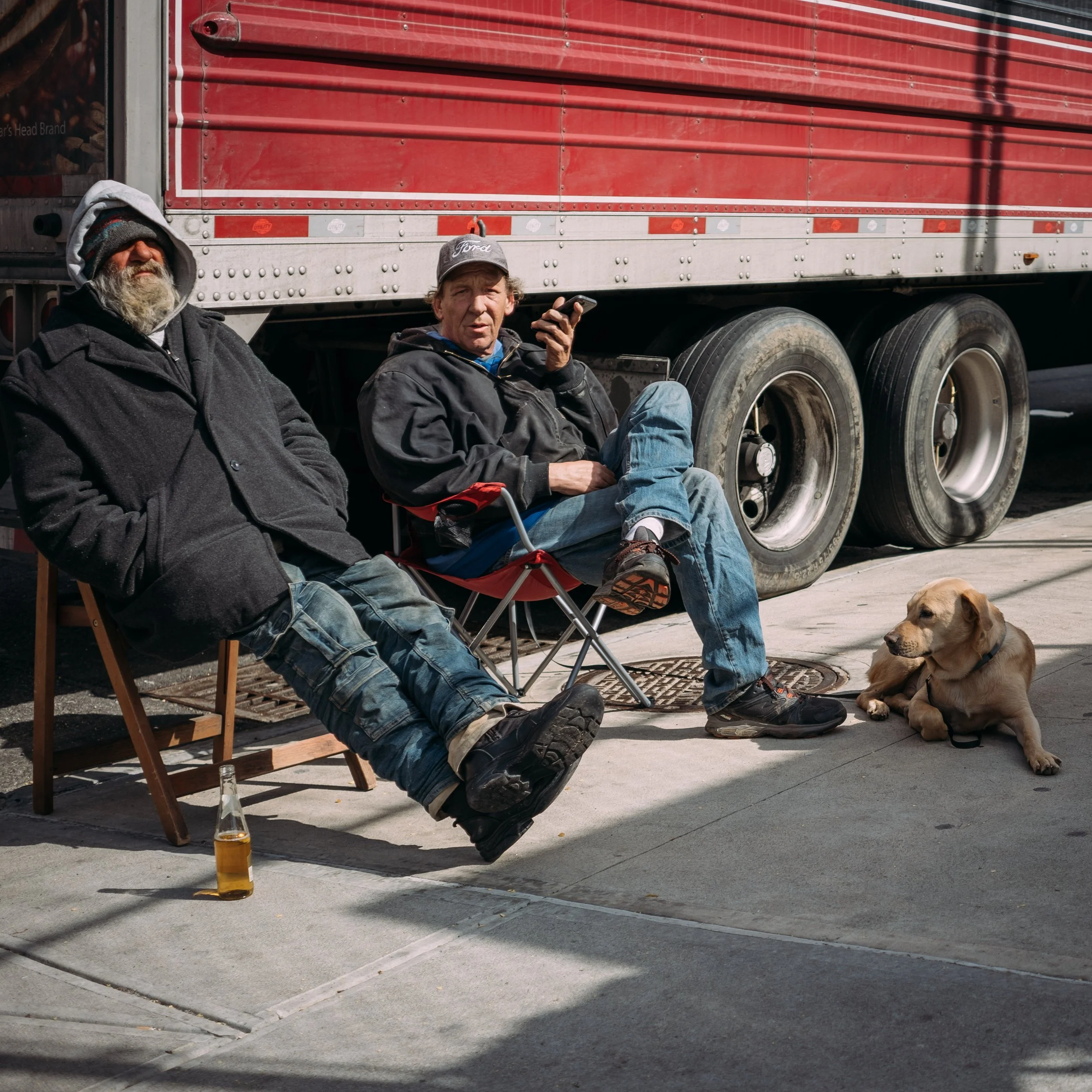 Two men sitting on the sidewalk near a red semi-truck, with a dog lying nearby. One man is wearing a cap and listening to a phone, the other has a beard and is wearing a hoodie and sunglasses. A beer bottle is on the ground.