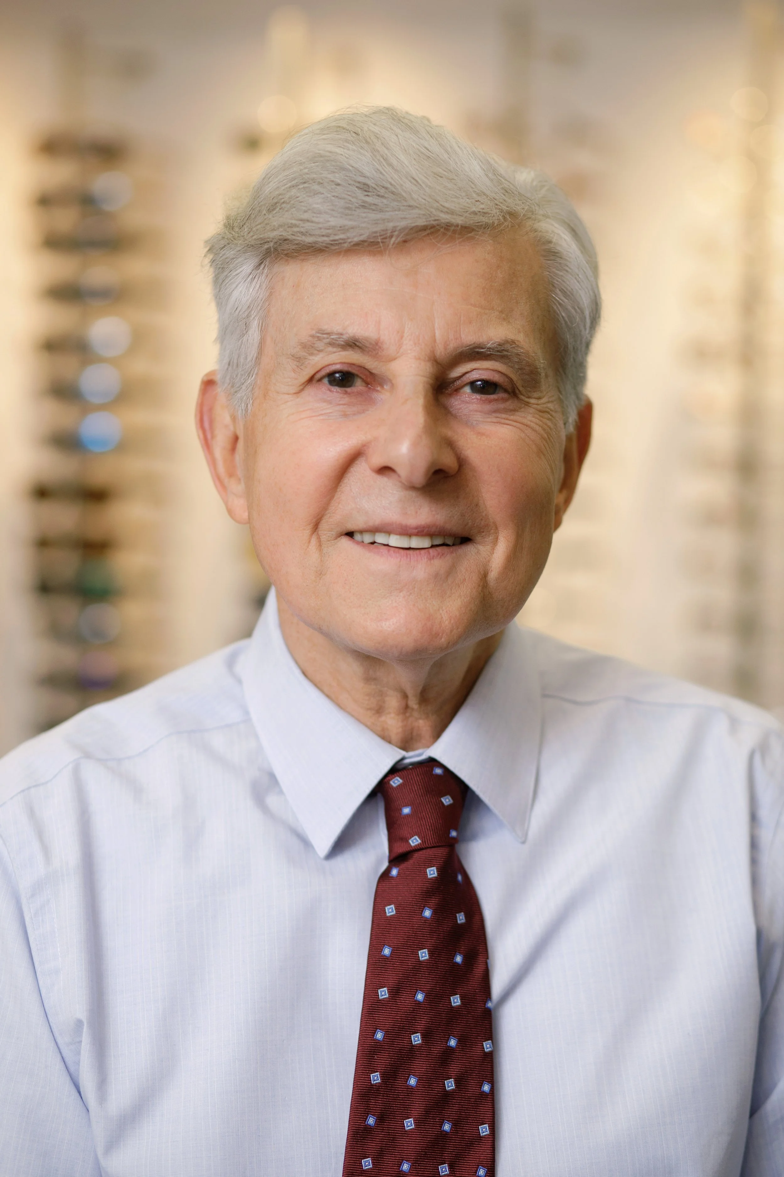 Headshot of an older man with silver hair wearing a light blue dress shirt and a maroon patterned tie, smiling in front of a blurred golden background.