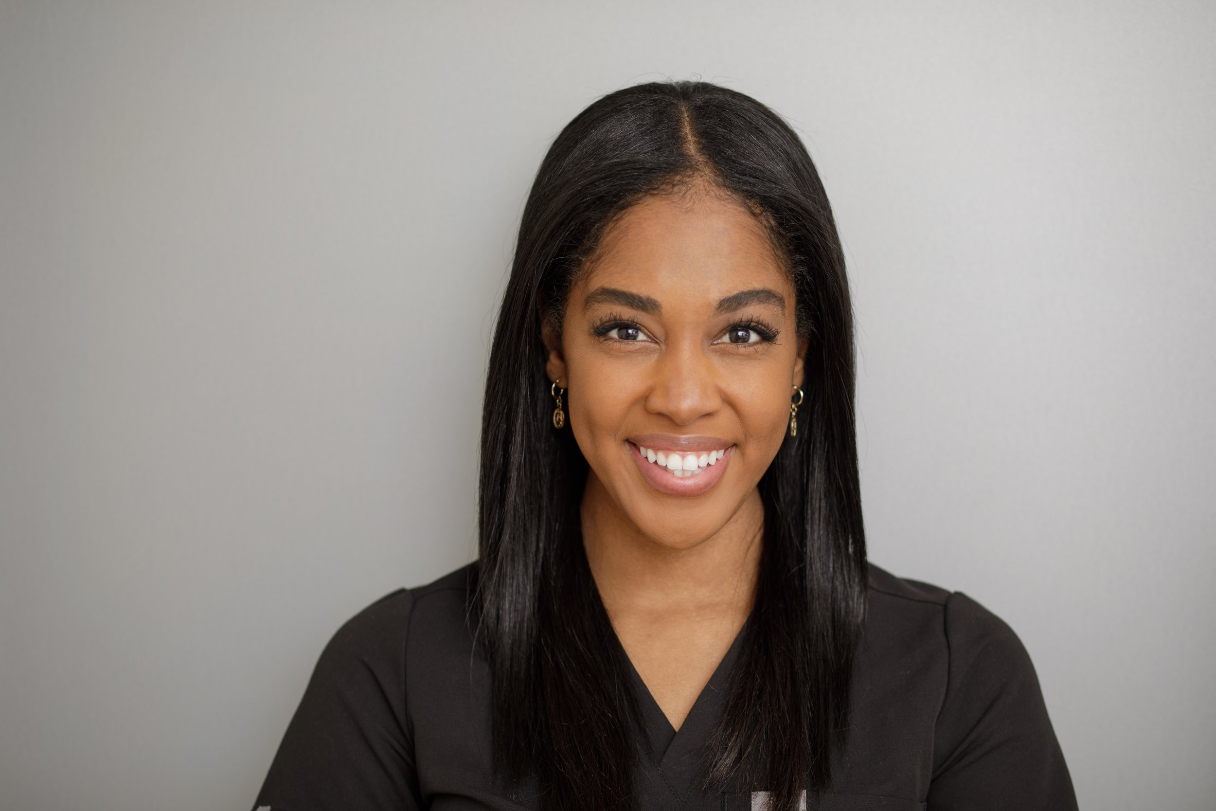 Smiling woman with long dark hair wearing earrings and a black top, standing against a plain light gray background.