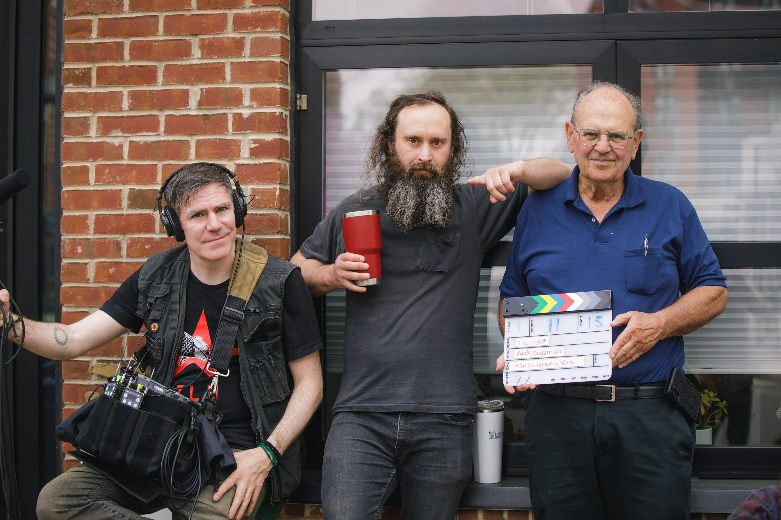 Three men standing in front of a brick building. The man on the right is holding a clapperboard with scripts written on it. The man in the middle has a long beard and is holding a red cup. The man on the left wears headphones and has a camera gear.