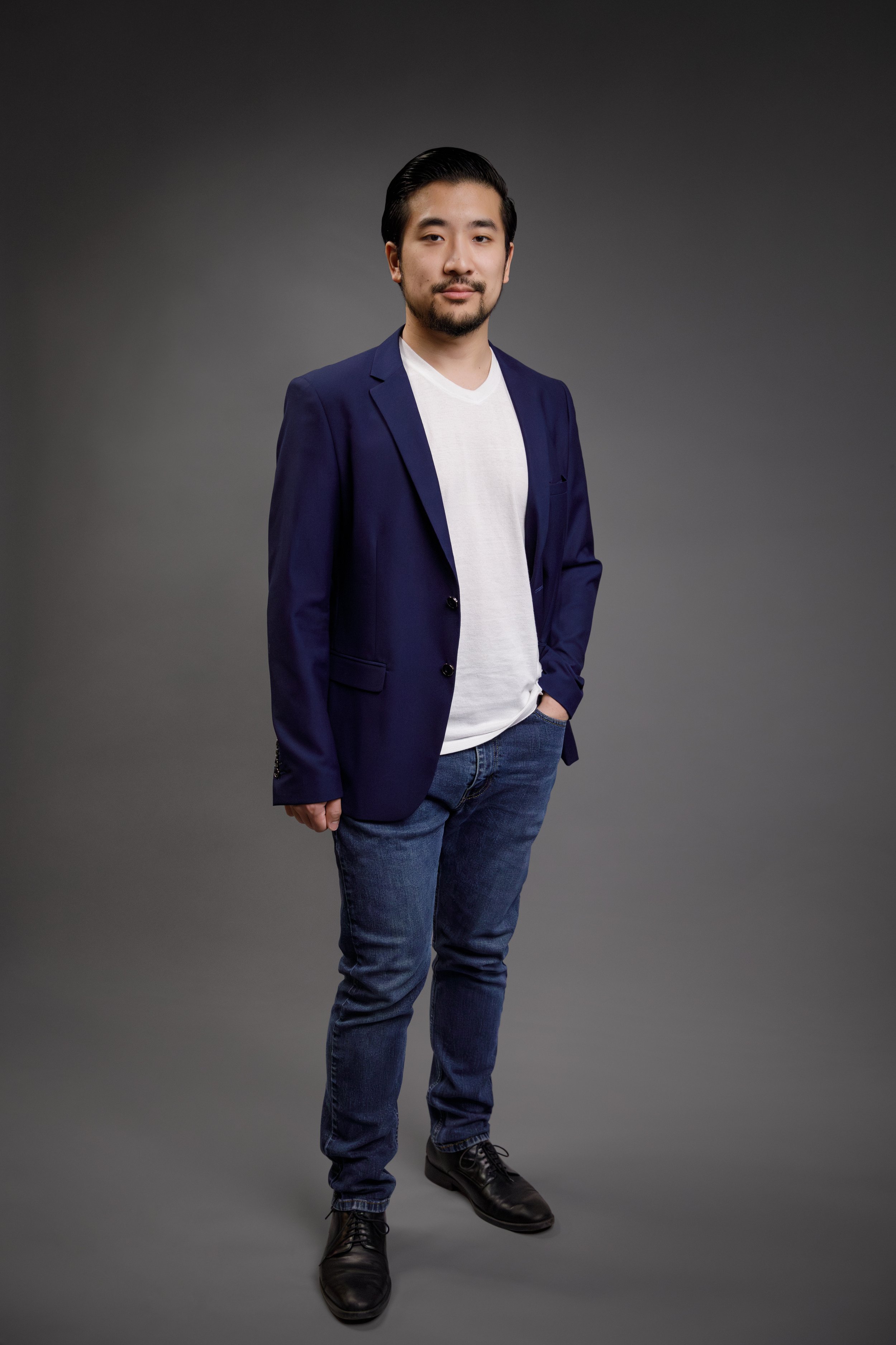 Portrait of a young man with dark hair and a beard, wearing a navy blazer over a white t-shirt, blue jeans, and black shoes, standing against a dark gray background.