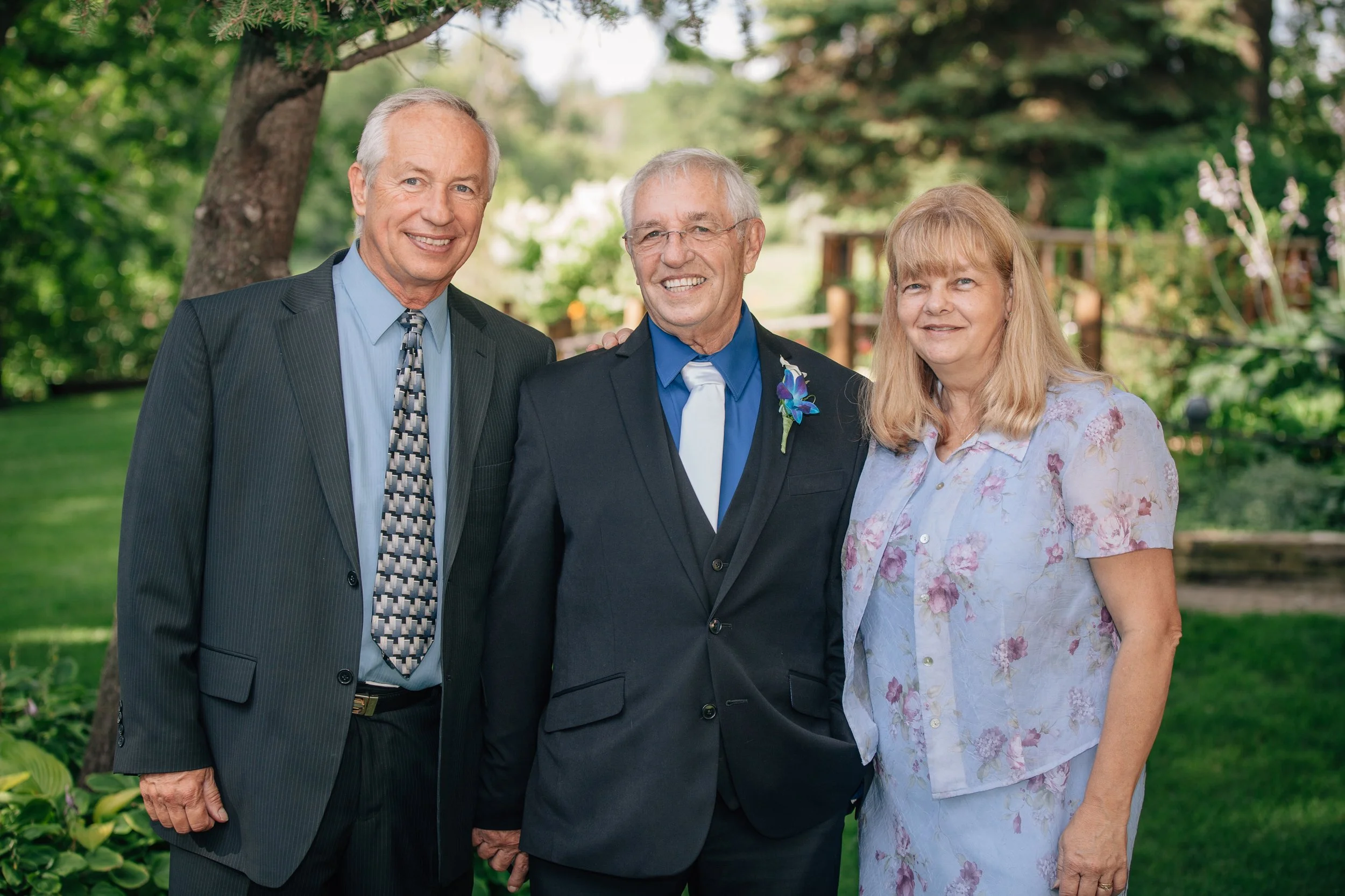 Three senior adults, two men and one woman, are standing outdoors in a green park, dressed in formal attire, smiling at the camera.