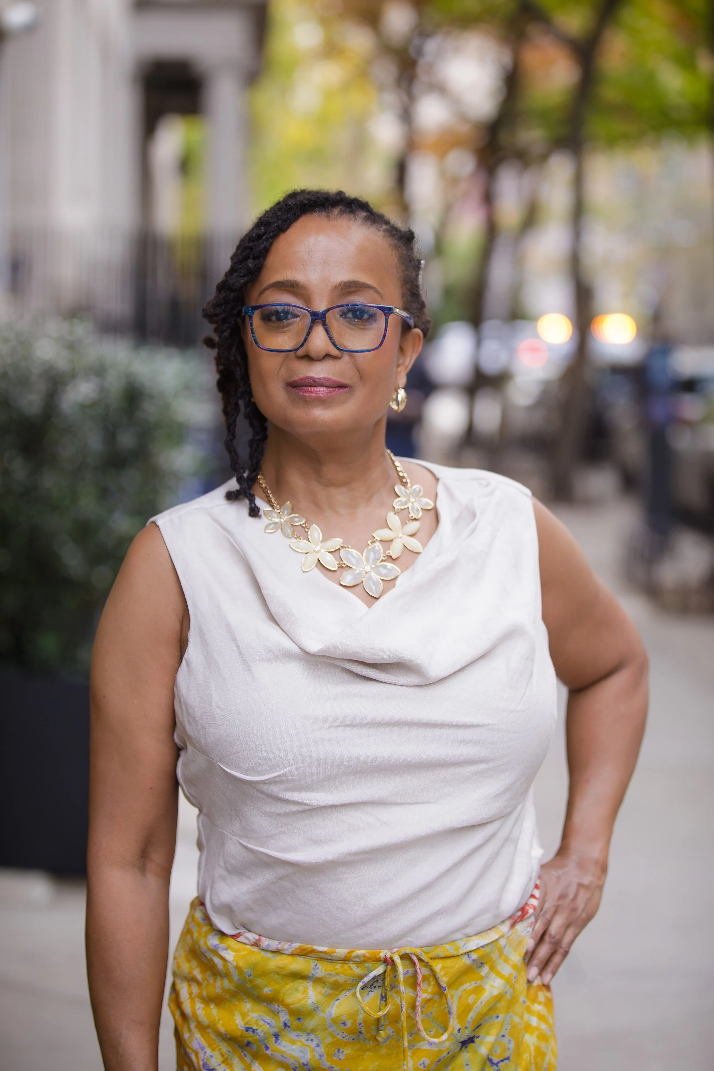A woman standing outdoors with trees and parked cars in the background. She has dark, styled hair, glasses, earrings, and a floral statement necklace. She's wearing a sleeveless white top and yellow patterned pants.