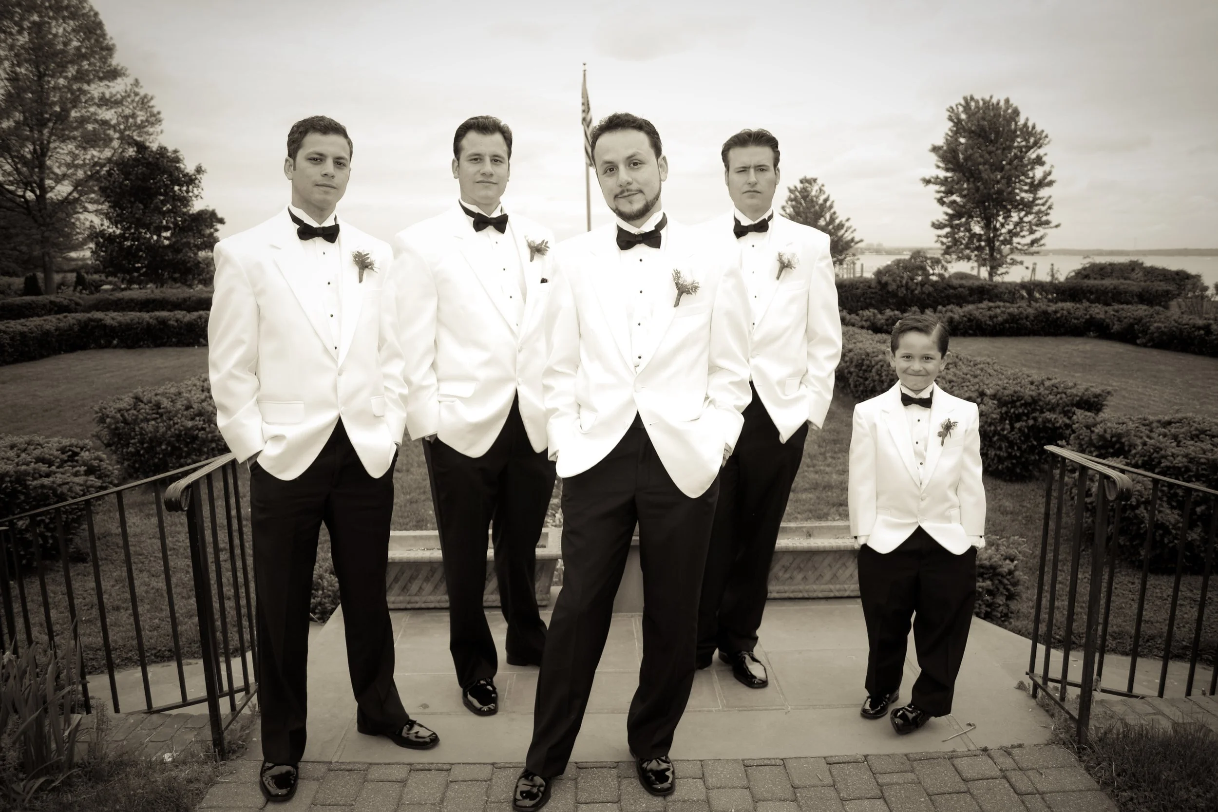 A group of five men and one boy in formal white tuxedos with black bow ties, standing outdoors on a pathway with a landscaped background. The scene is in black and white.