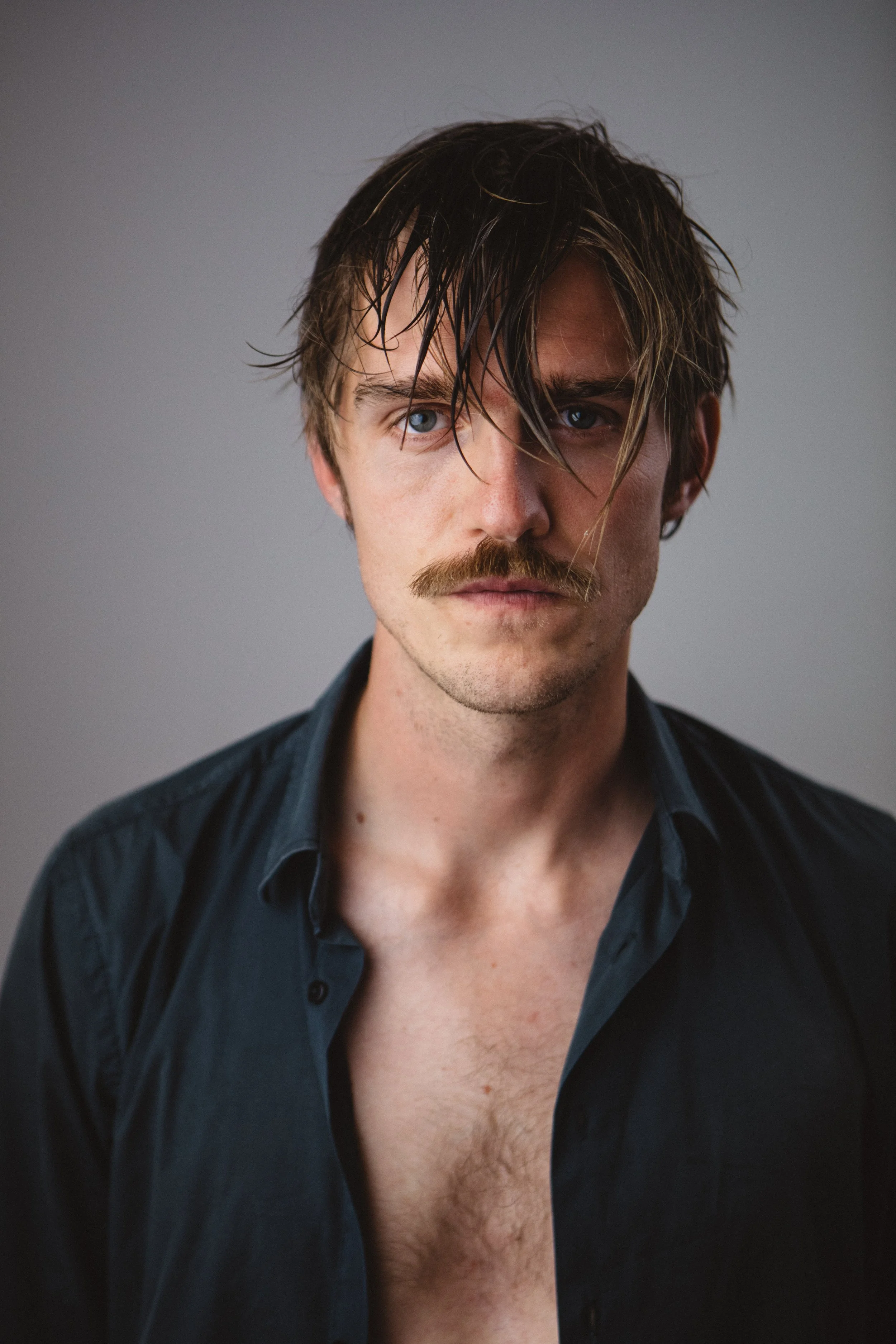 Close-up of a young man with wet, tousled brown hair, blue eyes, and a mustache, wearing a partially unbuttoned black shirt revealing chest hair, against a gray background.