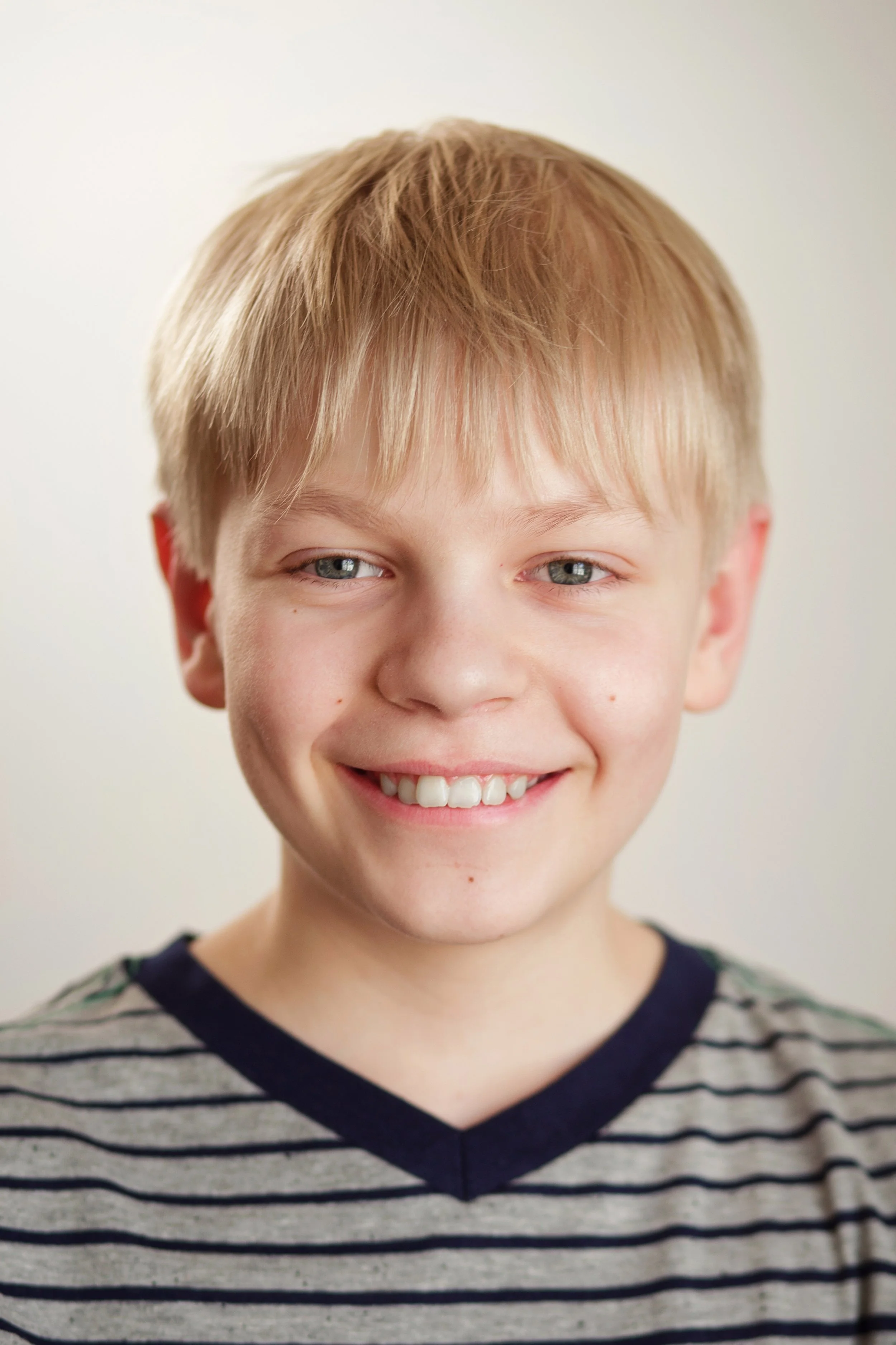 A smiling young boy with light blonde hair and blue eyes, wearing a gray and black striped shirt with a V-neck.