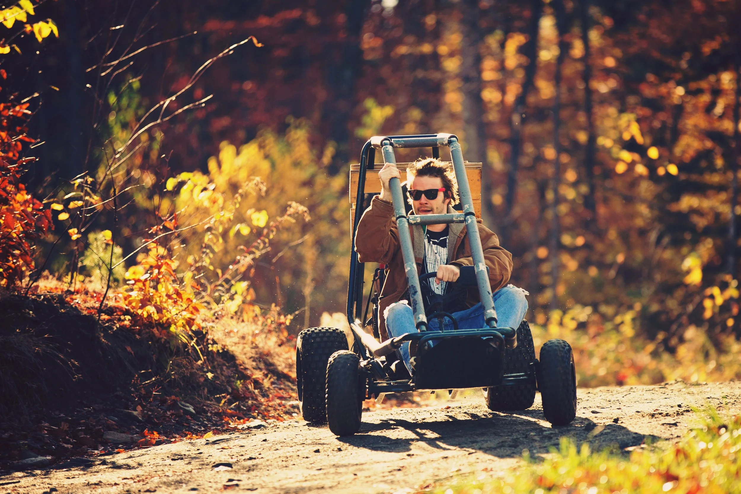A man with sunglasses riding a go-kart on an autumn forest trail surrounded by colorful fall foliage.