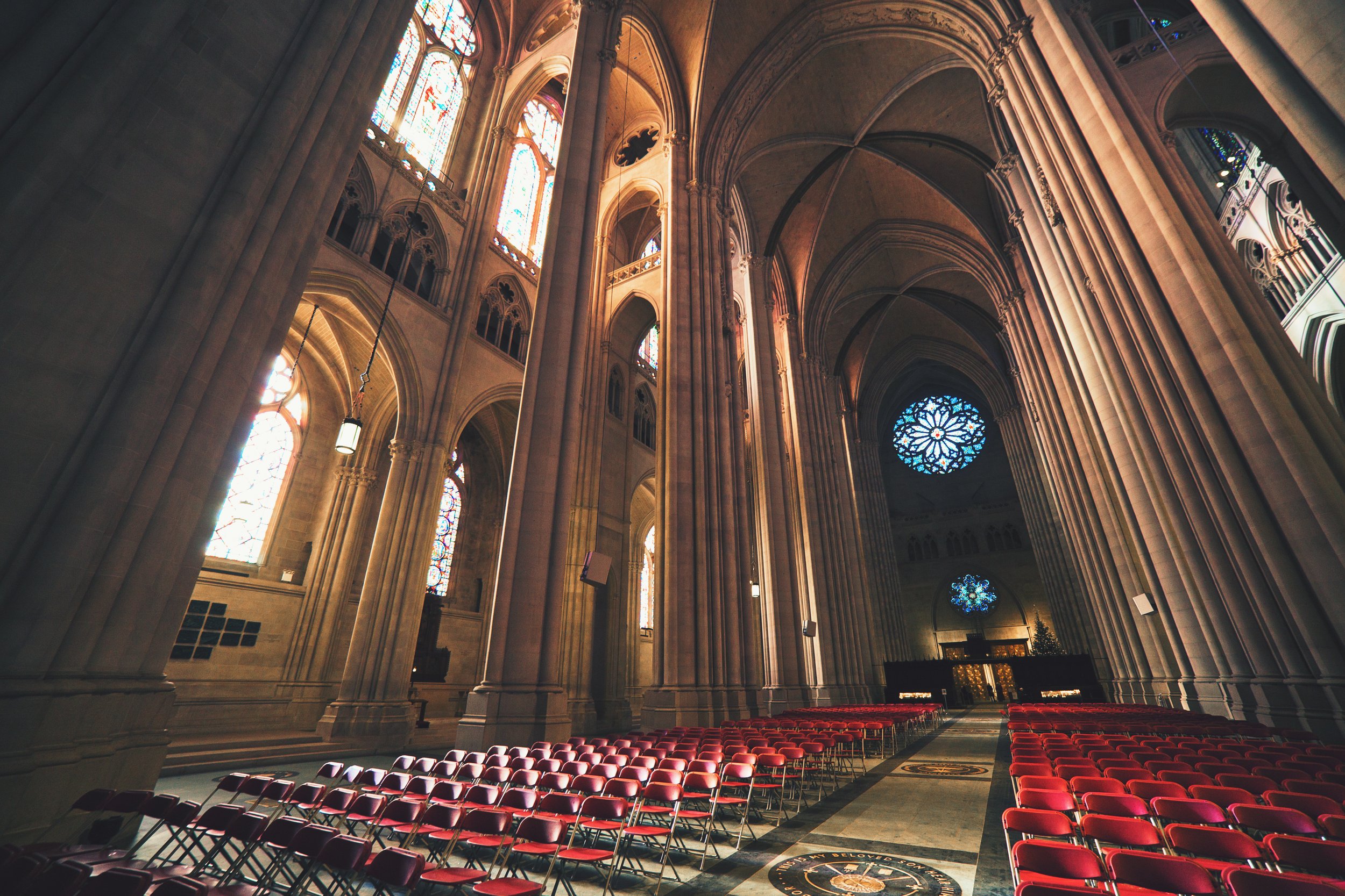Interior of a cathedral with high vaulted ceilings, large stained glass windows, and rows of red chairs arranged for a service or event.
