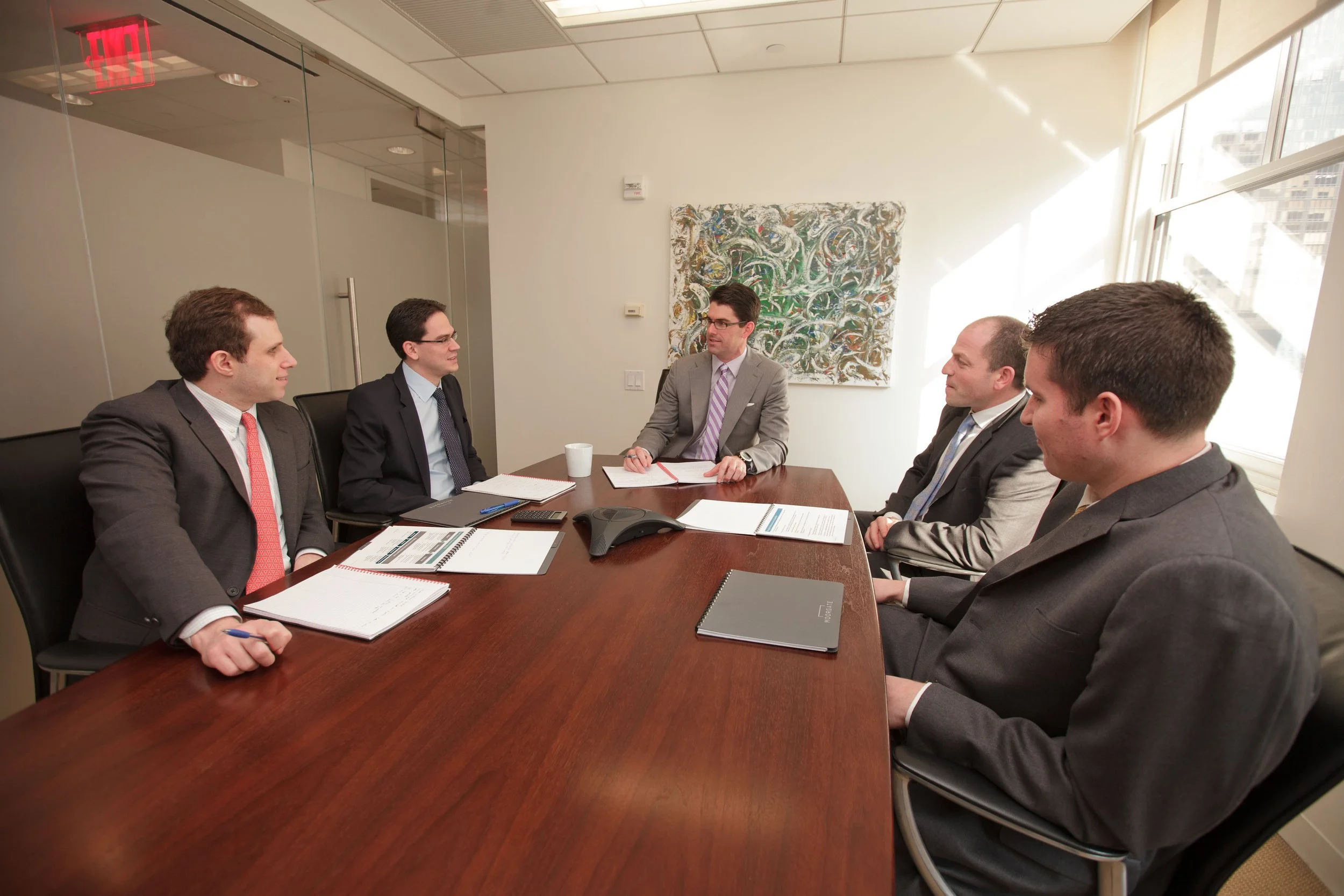 Five men in business suits having a meeting around a wooden conference table in a modern office with large windows and abstract artwork on the wall.