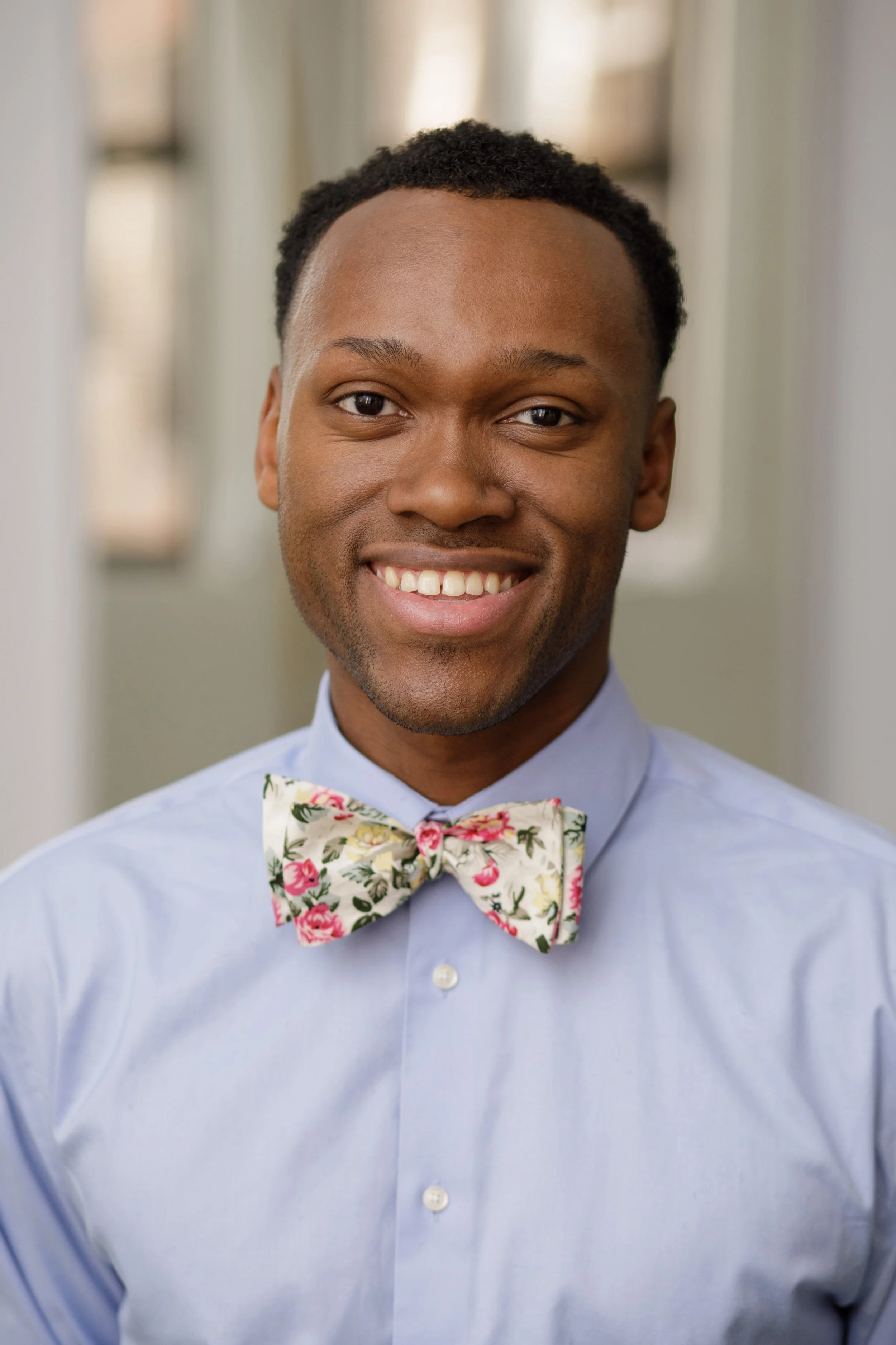 Close-up of a man smiling, dressed in a light blue shirt and a floral bow tie, standing in an indoor setting.