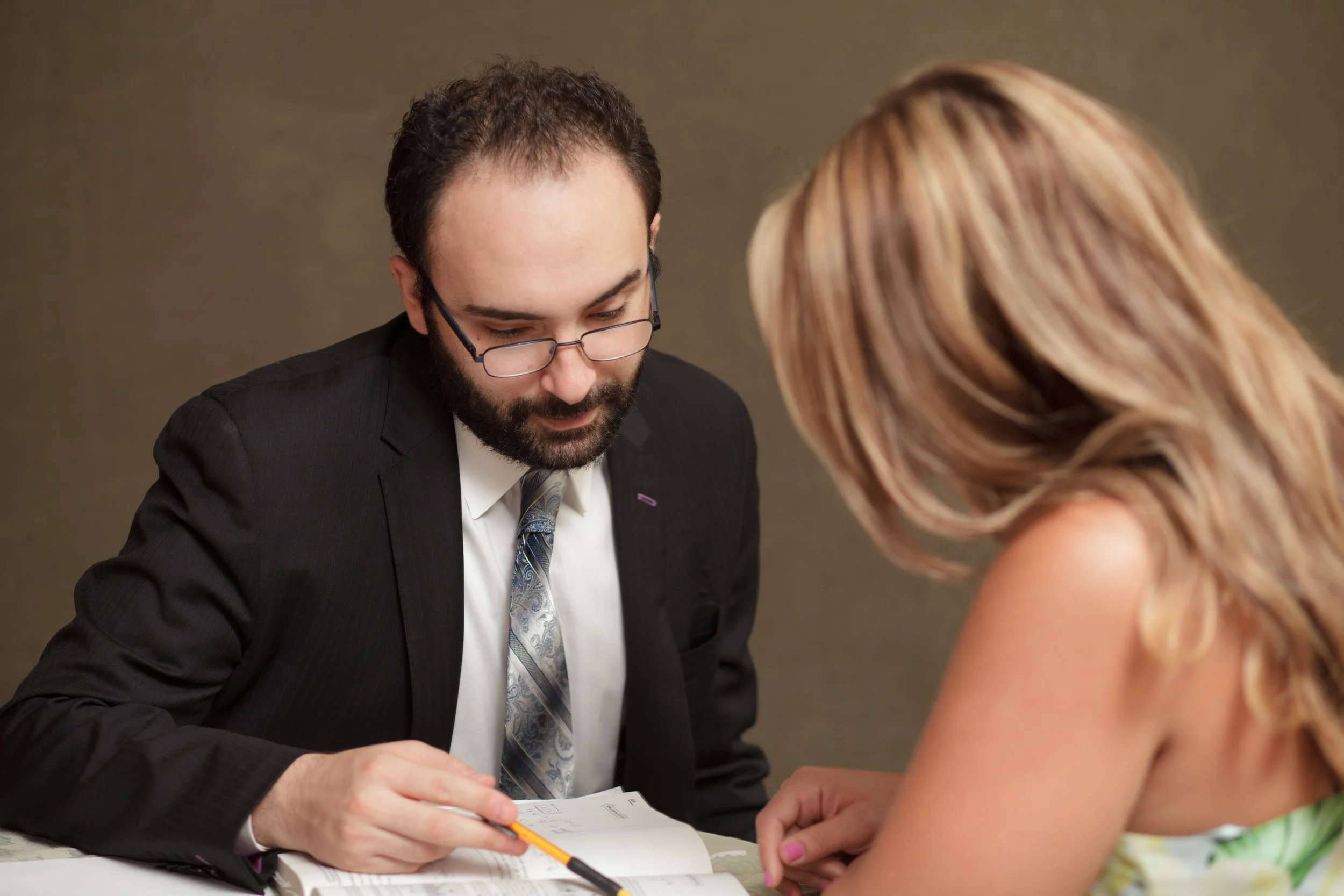 A man with glasses, a beard, and wearing a dark suit, white shirt, and a patterned tie, is looking down and writing in a notebook. A woman with blonde, wavy hair, dressed in colorful clothing, is also looking down at the notebook. They are sitting at