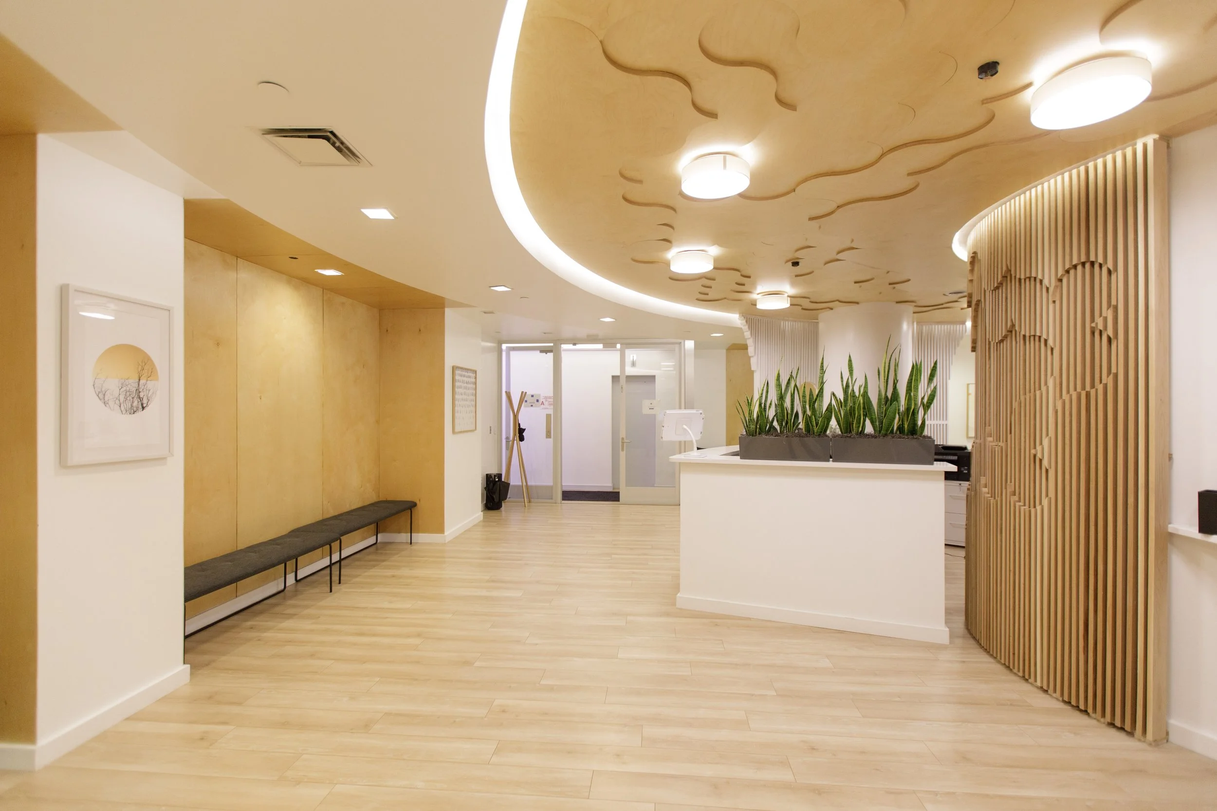 A modern interior space with wood flooring, light-colored walls, decorative ceiling lighting, and wooden wall accents, including a curved wooden partition and a planter with green plants, possibly a reception or waiting area.