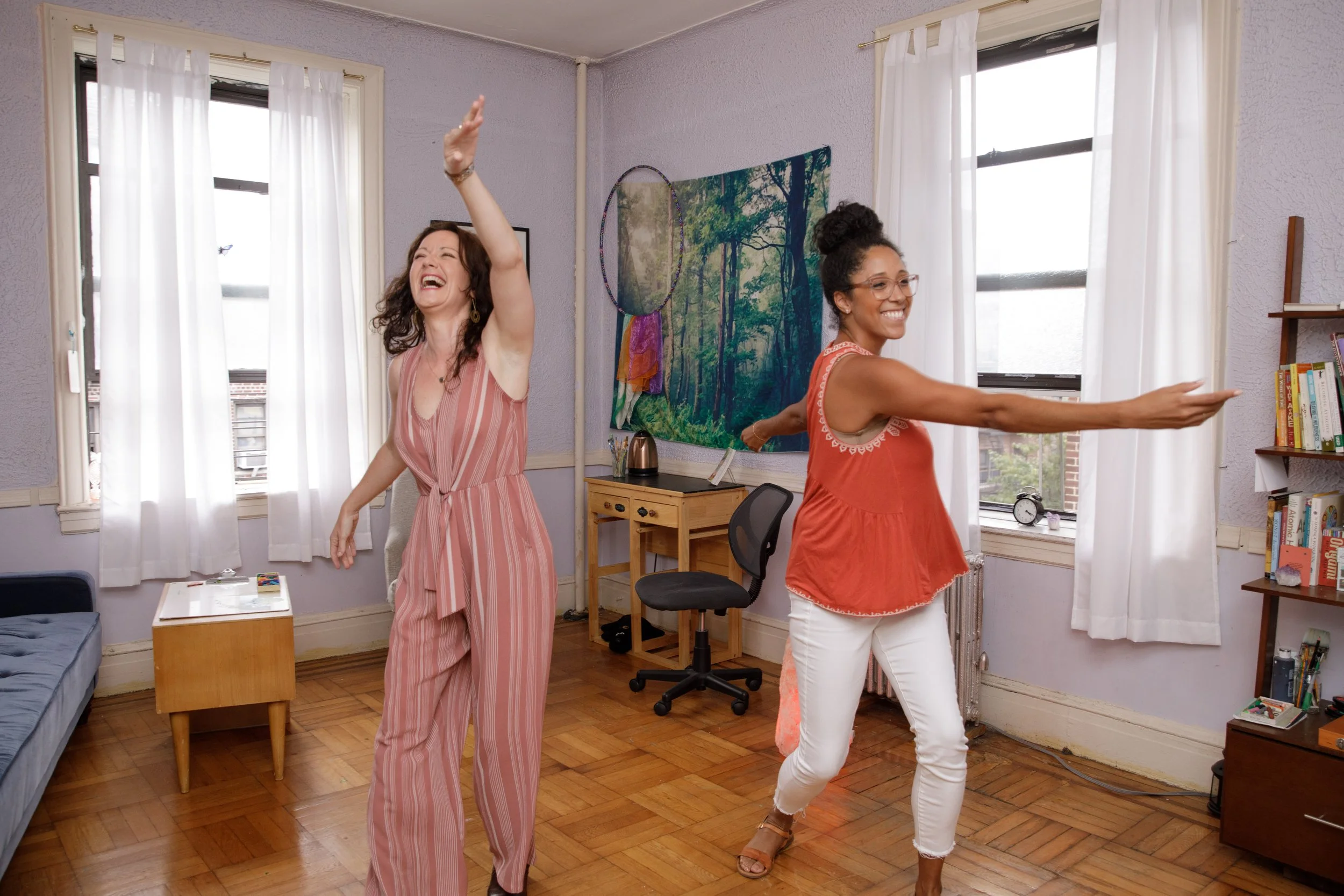 Two women dancing and laughing in a brightly lit room with large windows, white curtains, and wooden floors.