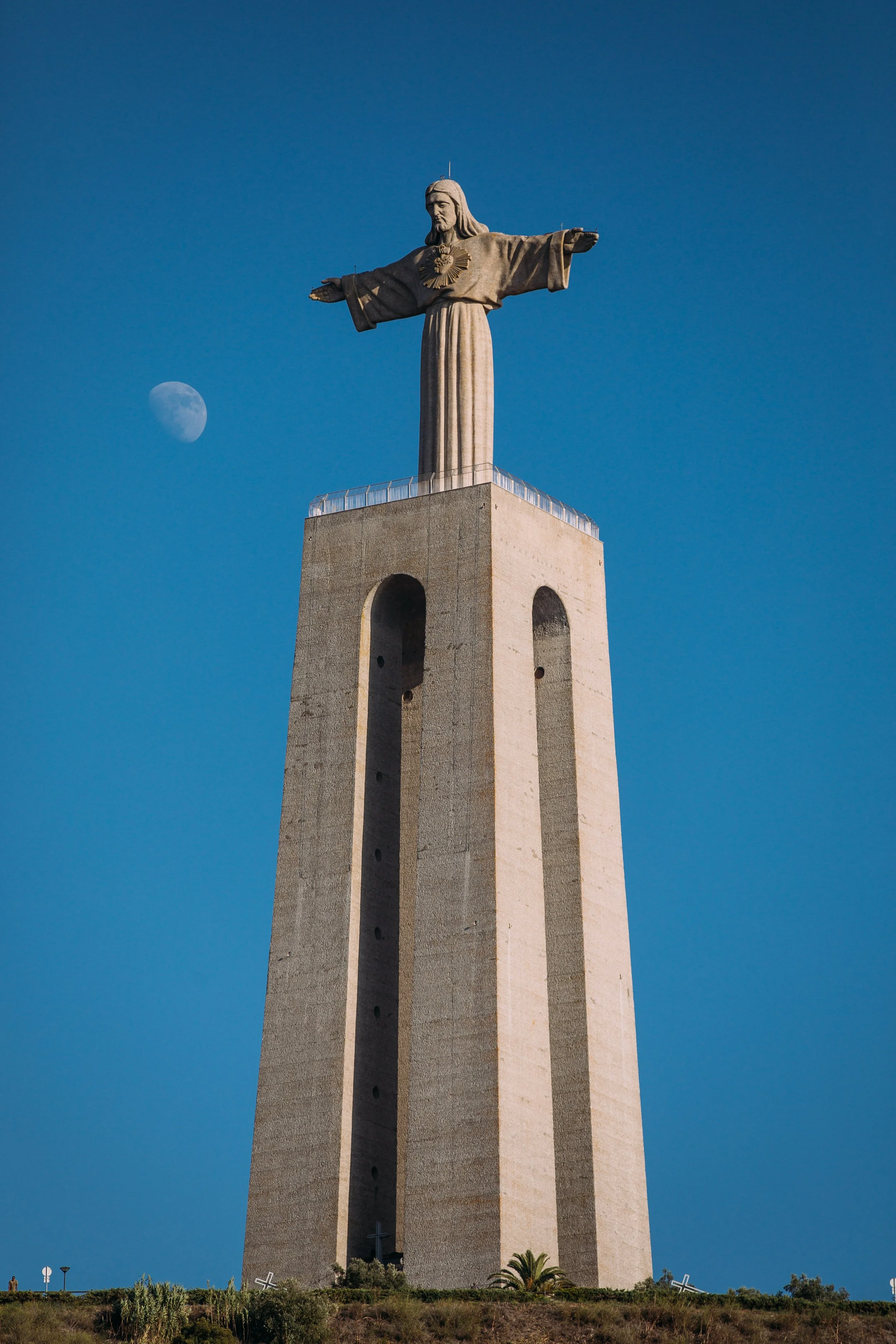 The Christ the King monument in Almada, Portugal, with a clear sky and the moon visible in the background.