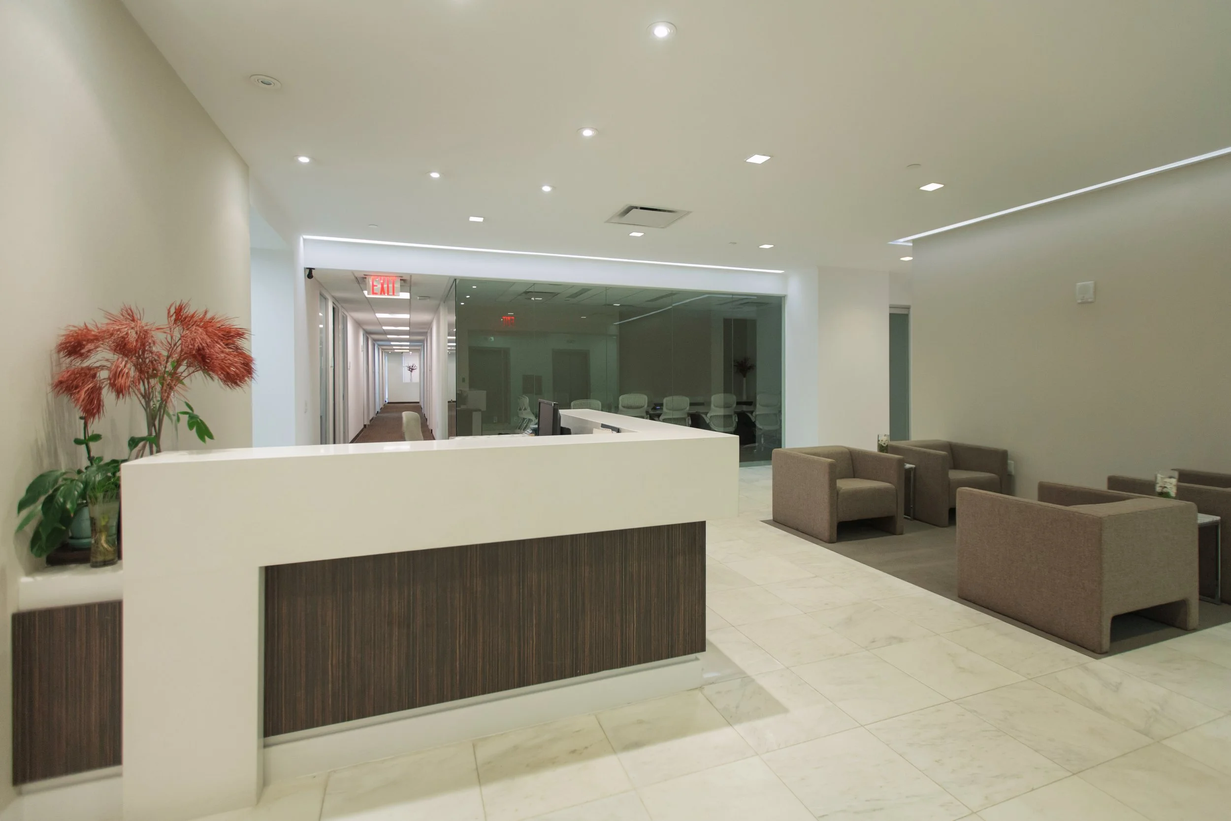 A modern office lobby with a curved reception desk, beige armchairs, a glass-enclosed conference room, and a hallway with chairs, white walls, and recessed ceiling lights.