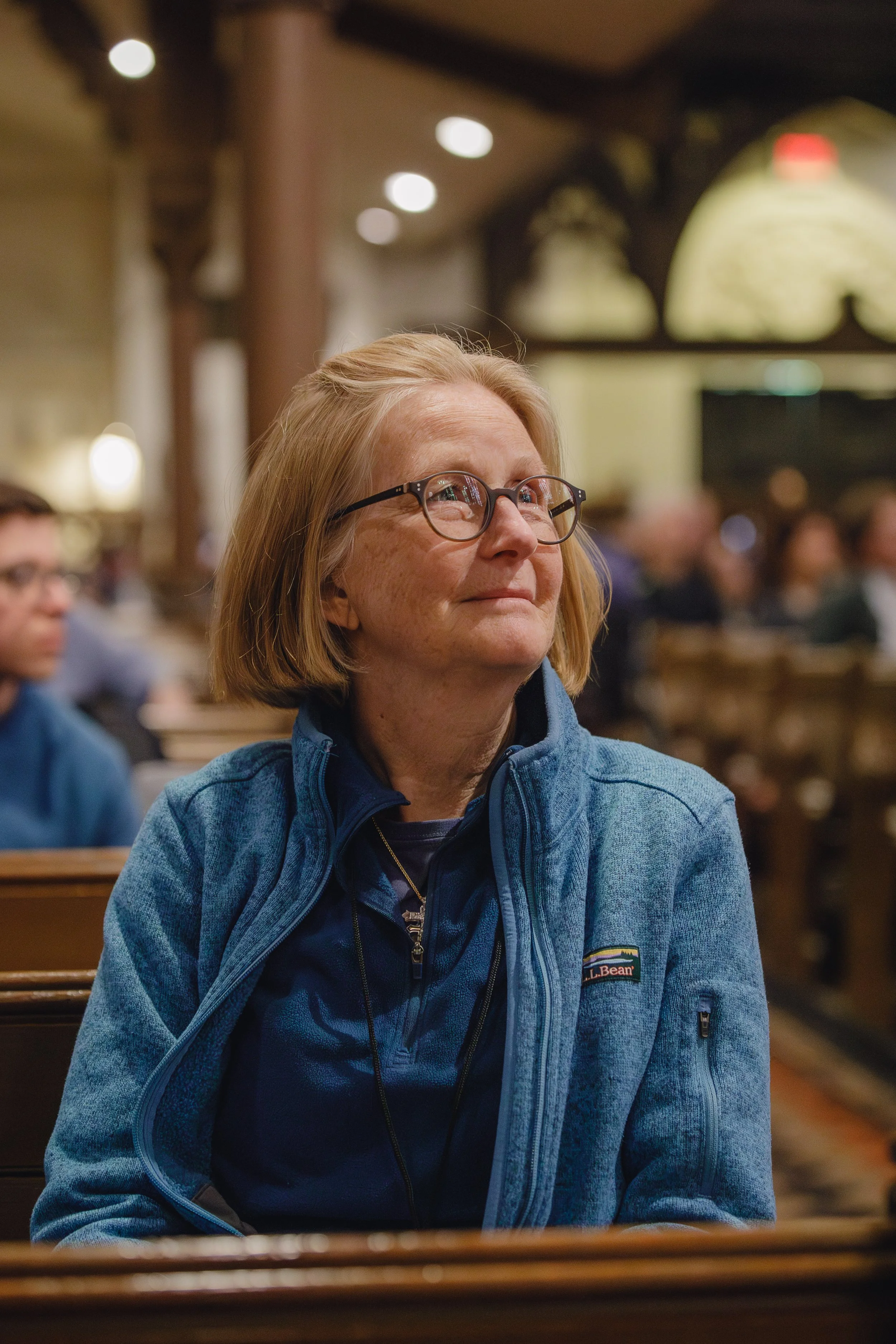 A woman with blonde hair and glasses wearing a blue jacket sitting in a restaurant with wood decor and blurred background of other people.
