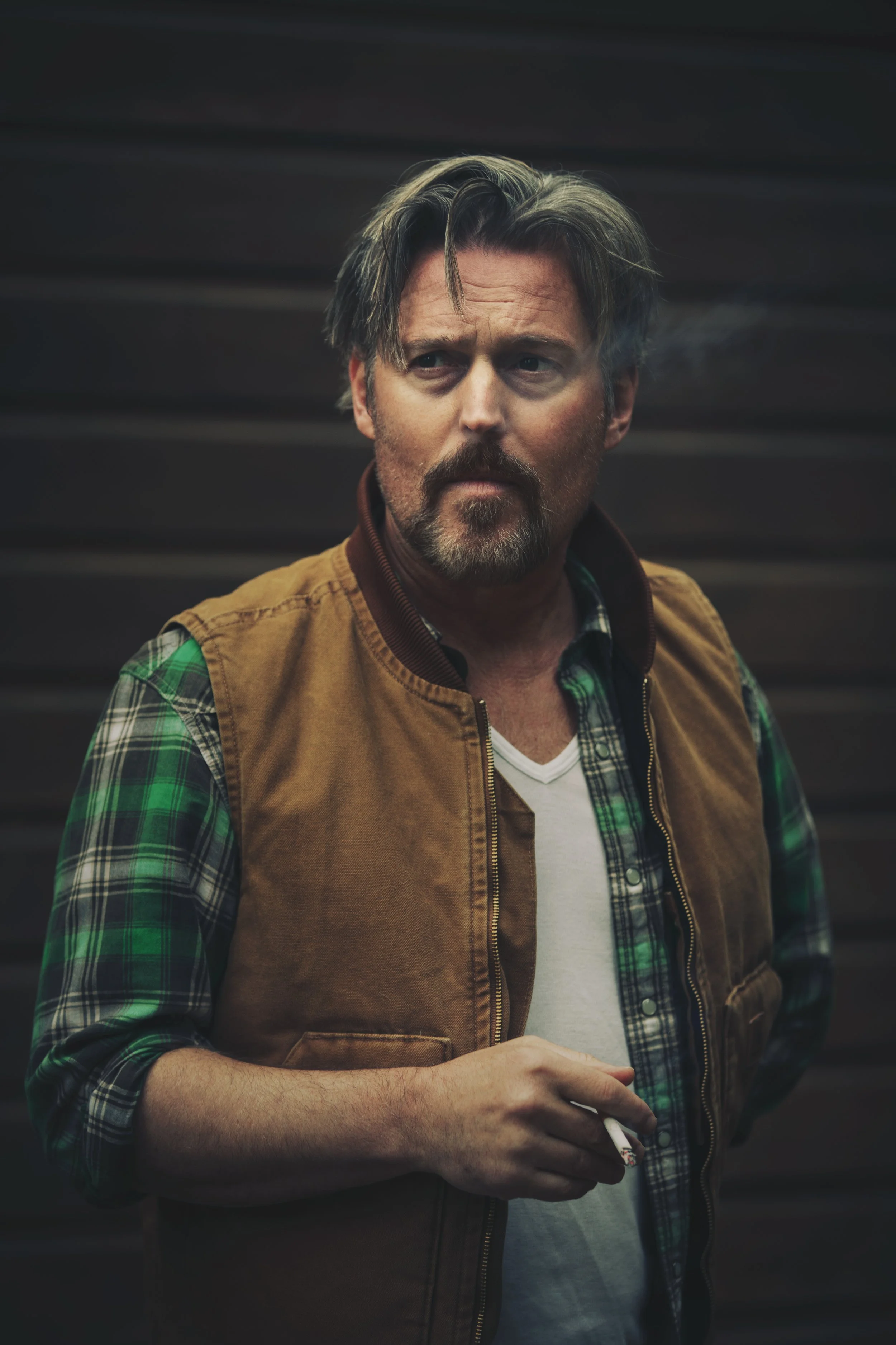 A man with a beard and unkempt hair wearing a brown vest over a green and black plaid shirt, holding a lit cigarette.