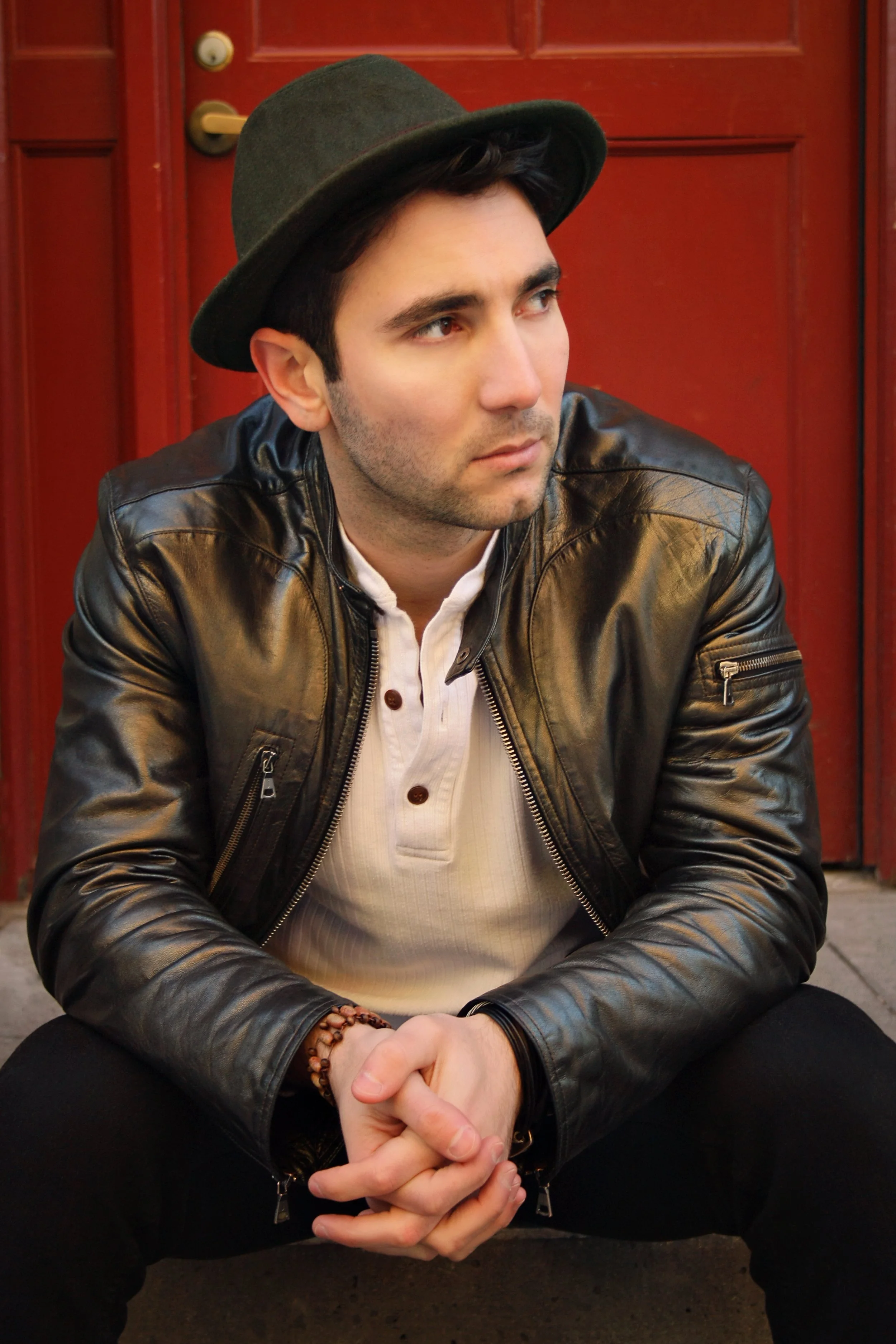 A young man wearing a gray hat, black leather jacket, and white shirt sitting against a red door with their hands clasped.