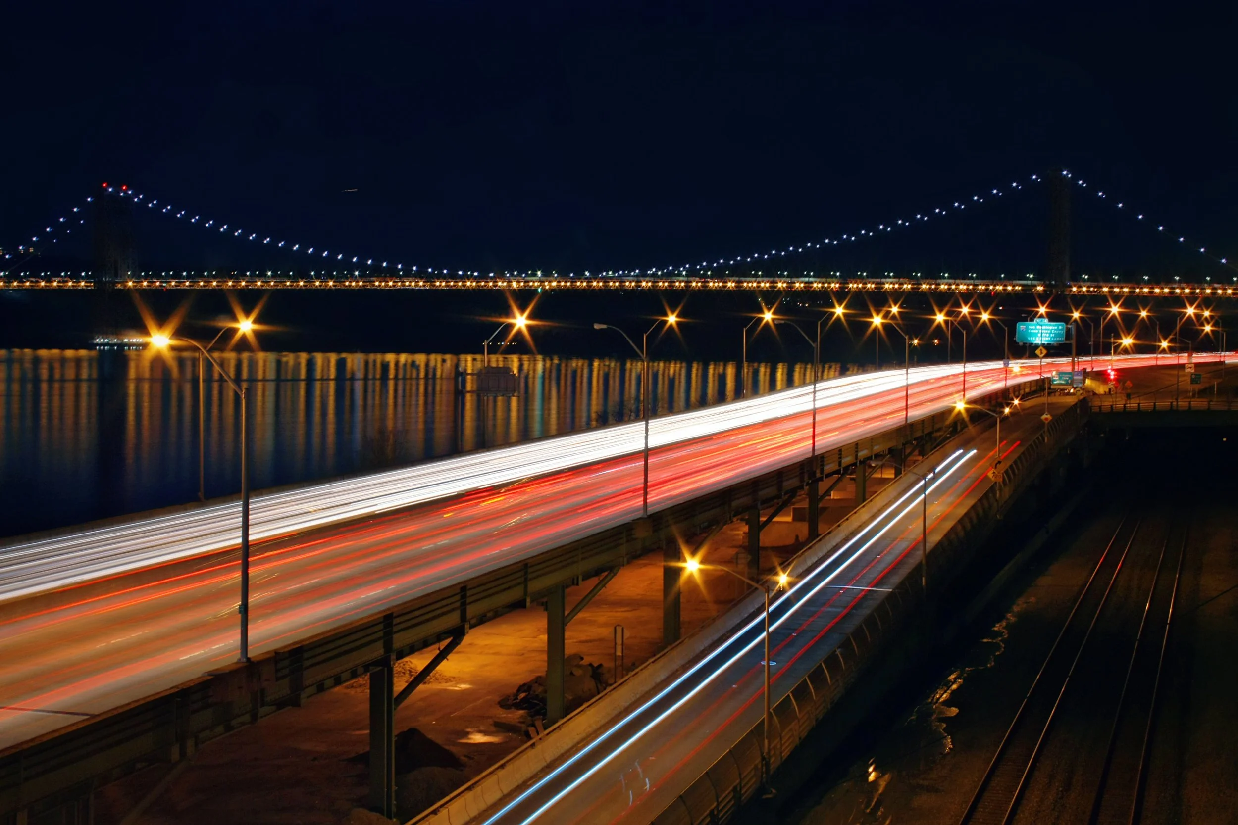 Nighttime view of a bridge illuminated with lights, with car light streaks on an adjacent road and reflections on the water below.