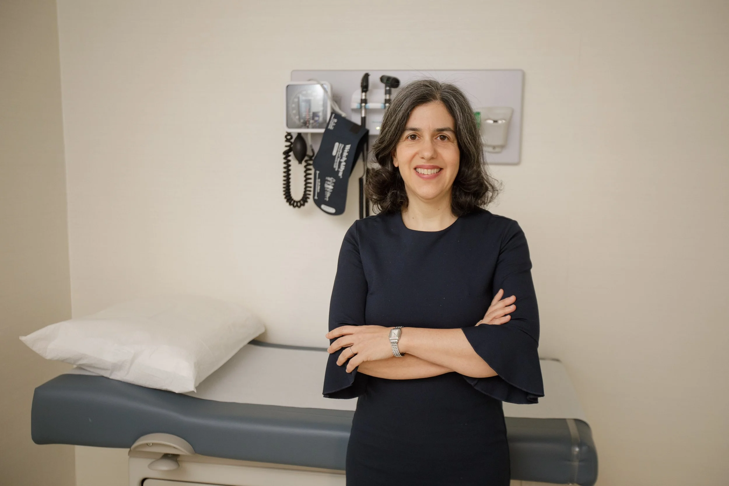 A woman with dark, grayish hair smiling, standing arms crossed in a medical examination room with a pillow on an examination bed and medical equipment on the wall behind her.
