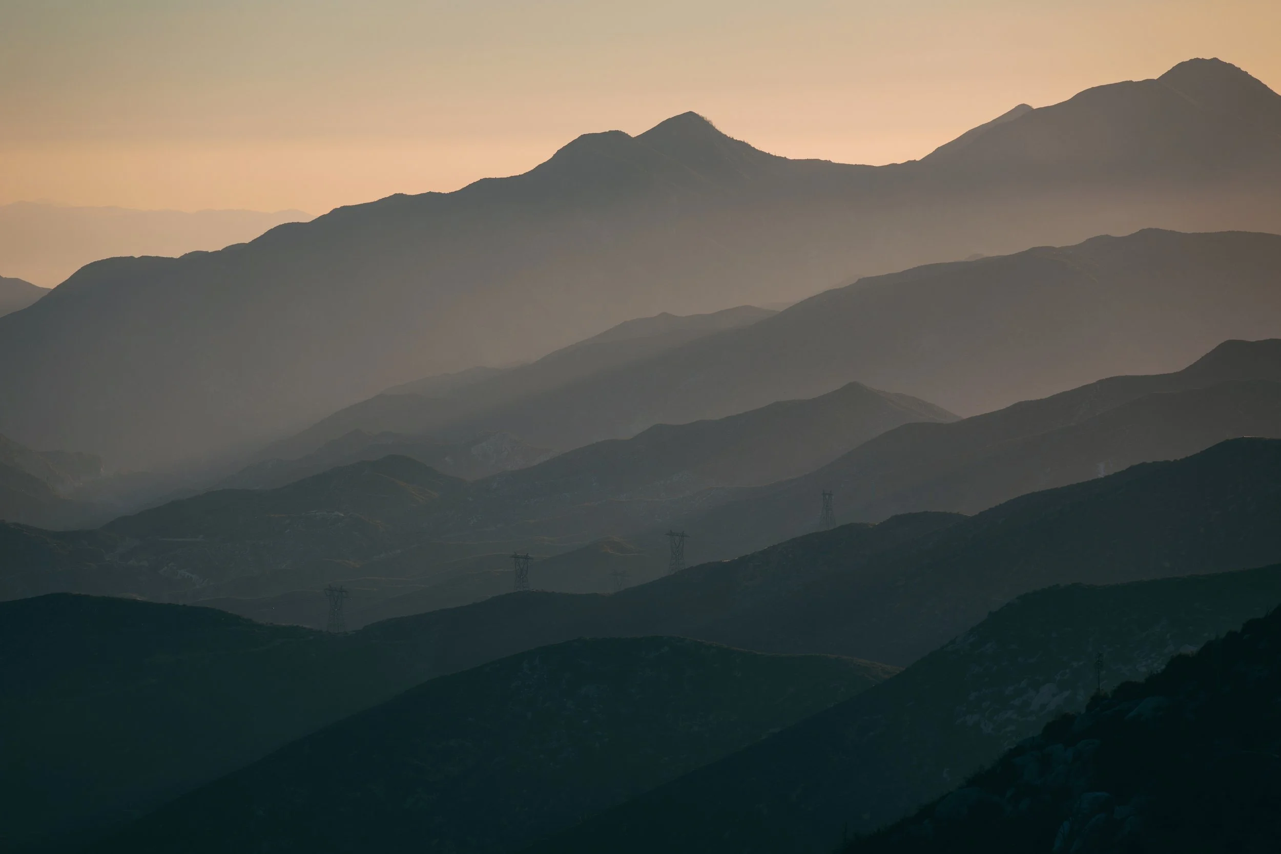 Photo of layered mountain ranges at sunset with an orange-pink sky and electricity pylons visible in the foreground.