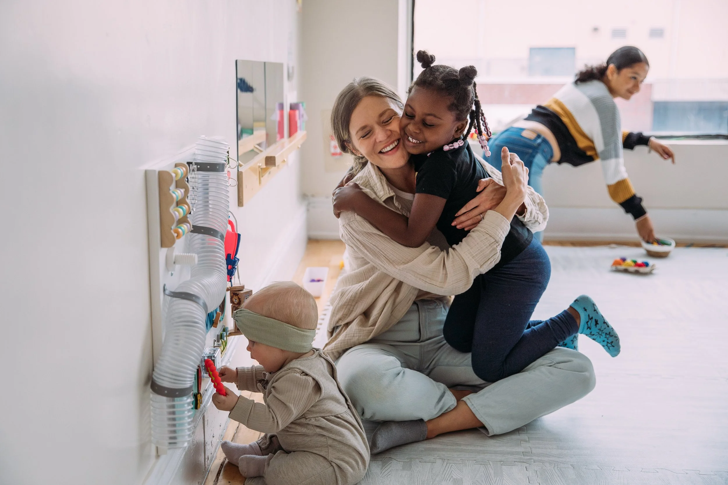 A woman with brown hair hugging a young girl with dark skin and hair styled in bantu knots, both smiling, with children playing in the background in a room with natural light, white walls, and colorful toys on the floor.