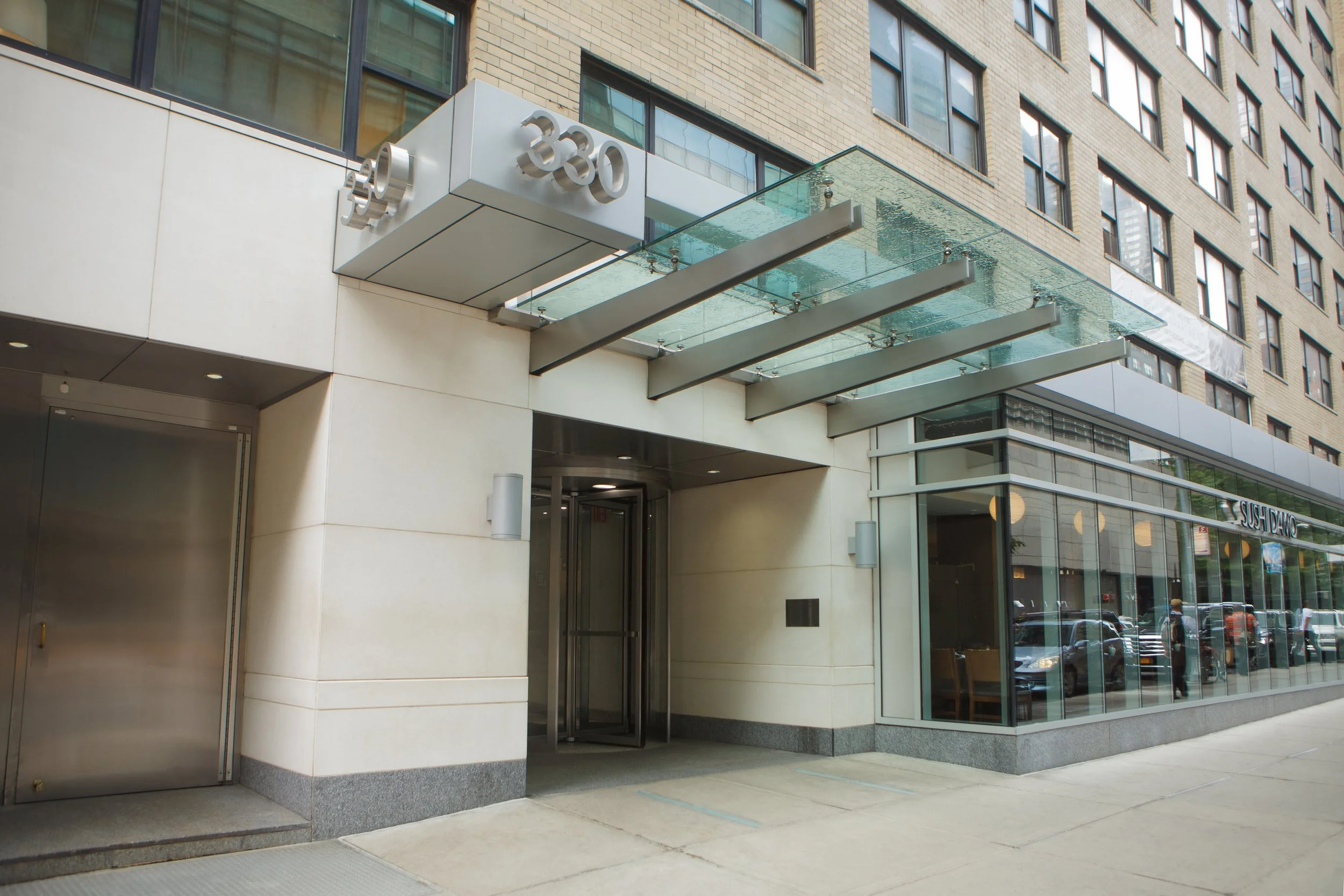 Street-level view of a modern building with a glass and metal entrance canopy, a sign displaying the number 330, glass windows, and a corner storefront.