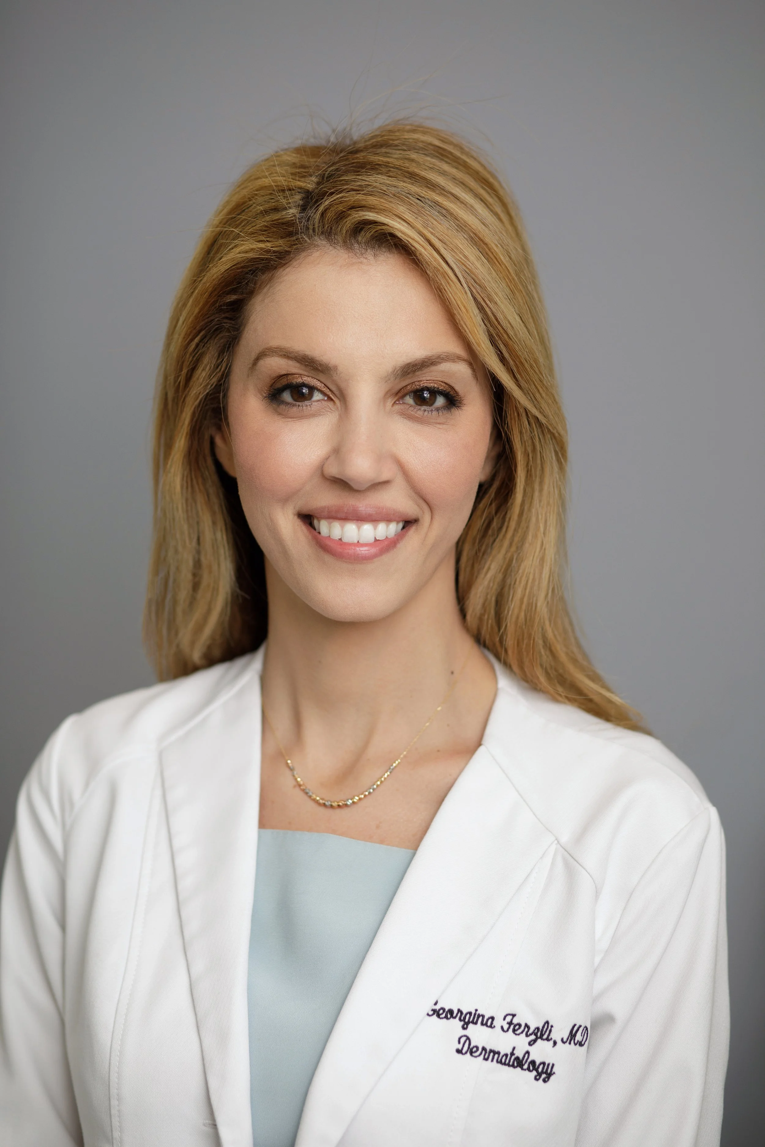 Photo of a smiling woman with blonde hair, wearing a white coat with embroidered name and profession, and a light blue top. She is also wearing a gold necklace. Gray background.
