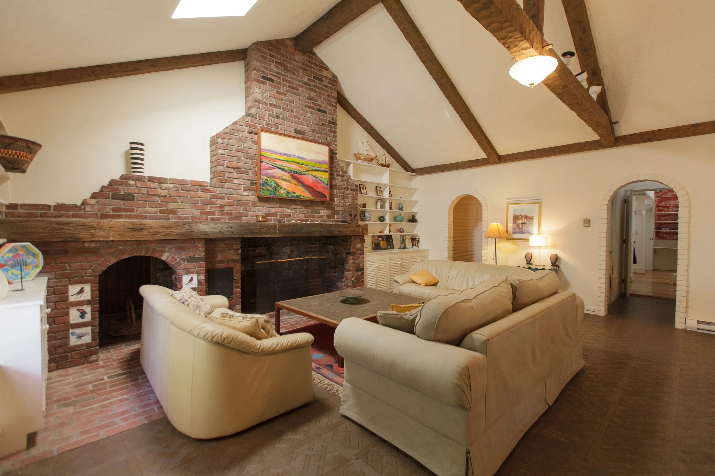 A living room with a brick fireplace, a painting above it, beige sofas, a wooden coffee table, bookshelves, lamps, and wooden ceiling beams.