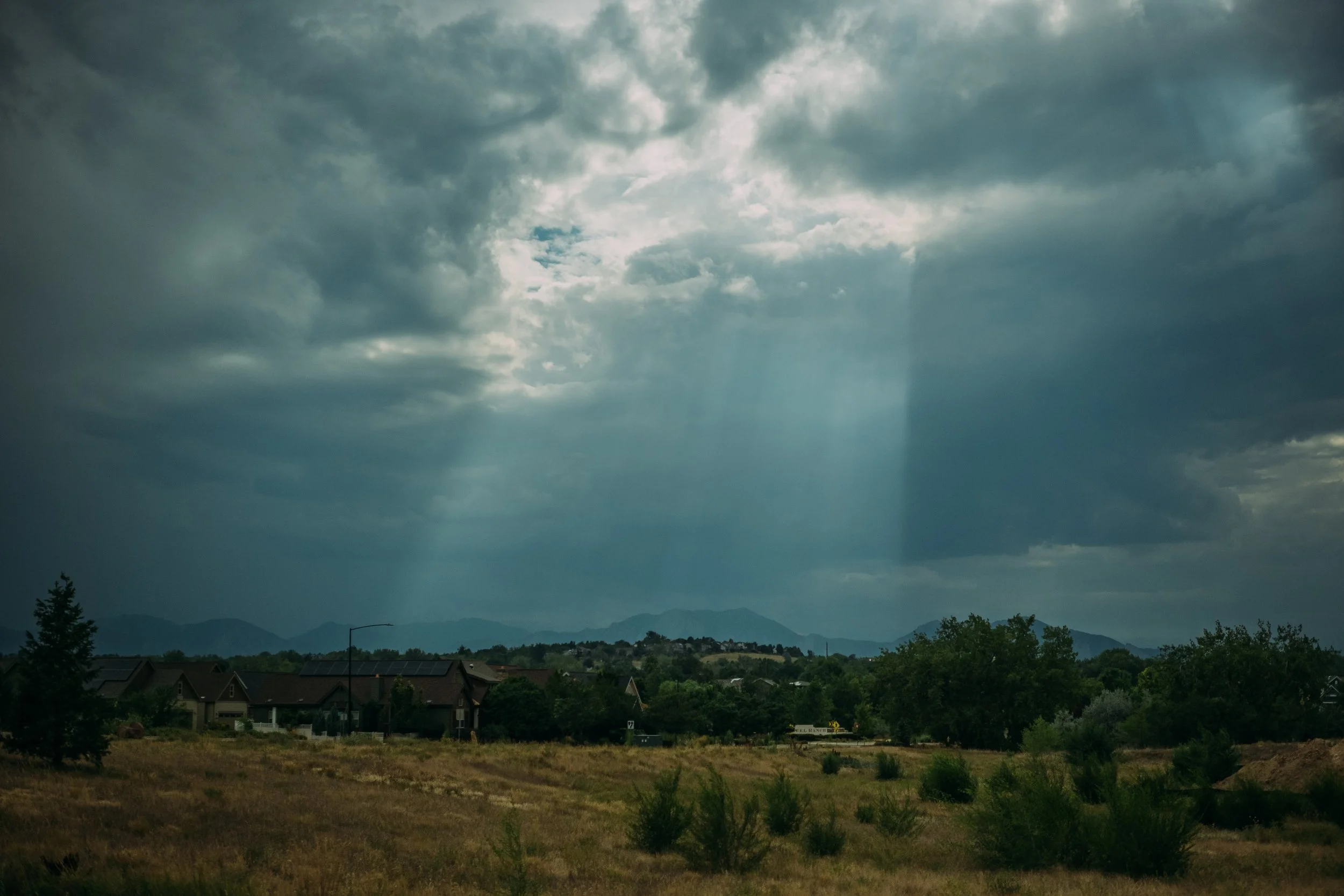 A landscape view of a suburban neighborhood with houses, trees, and mountains in the background under a cloudy sky with some sunlight breaking through.