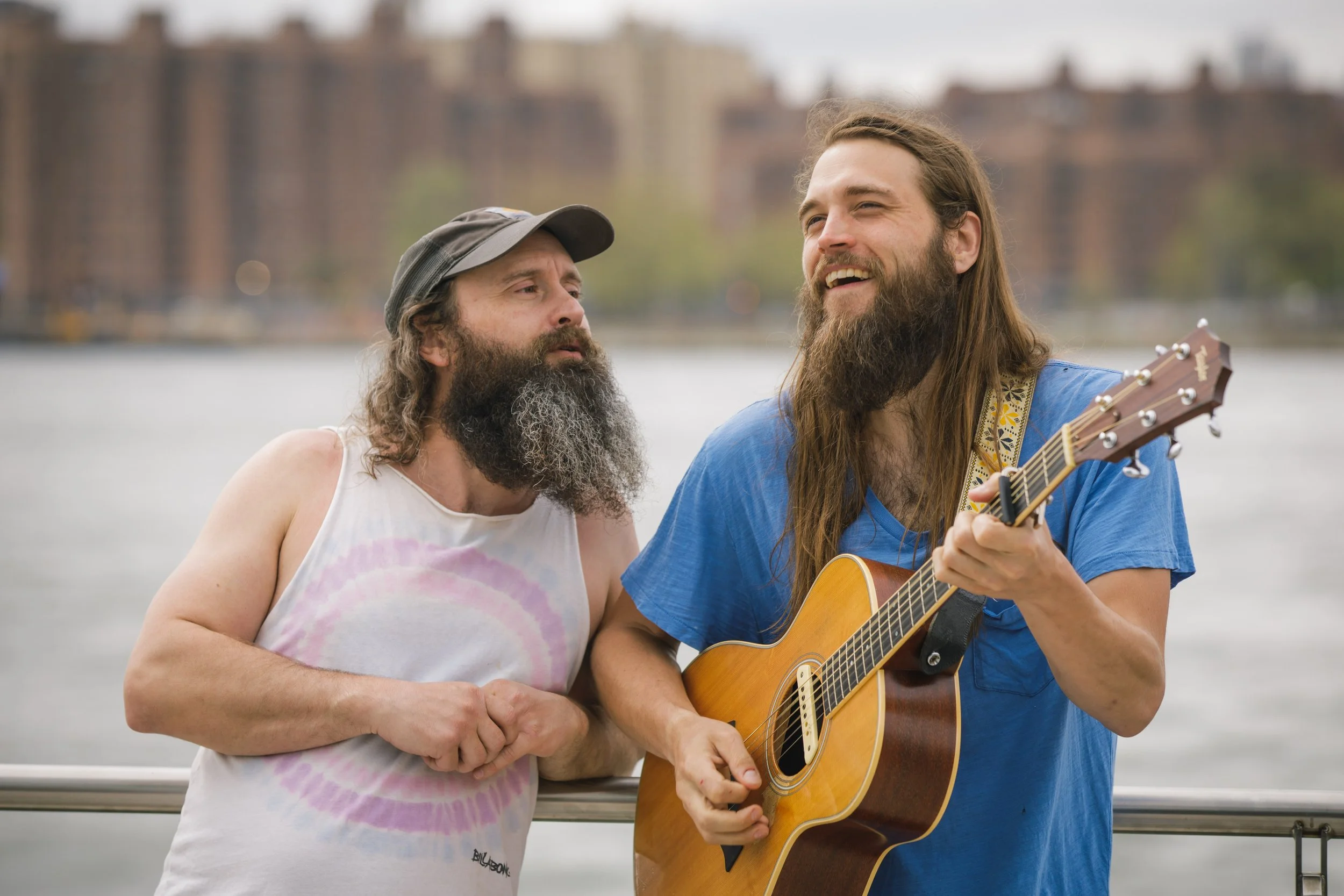 Two men with long hair and beards playing guitar and singing together outdoors near a body of water, with a cityscape in the background.