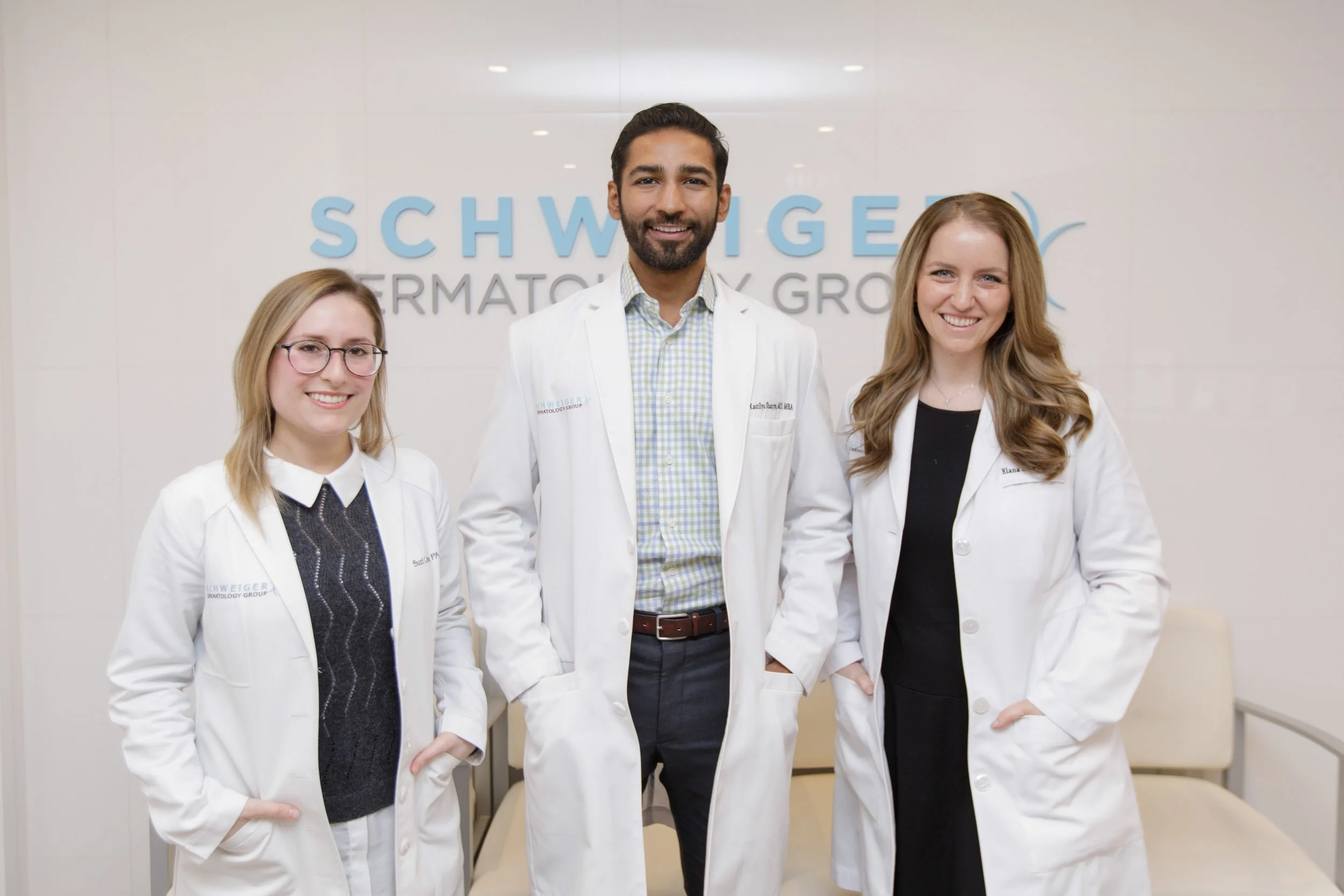 Four medical professionals in white coats standing in front of a white wall with the Schweiger Dermatology Group logo. They are smiling and looking at the camera.