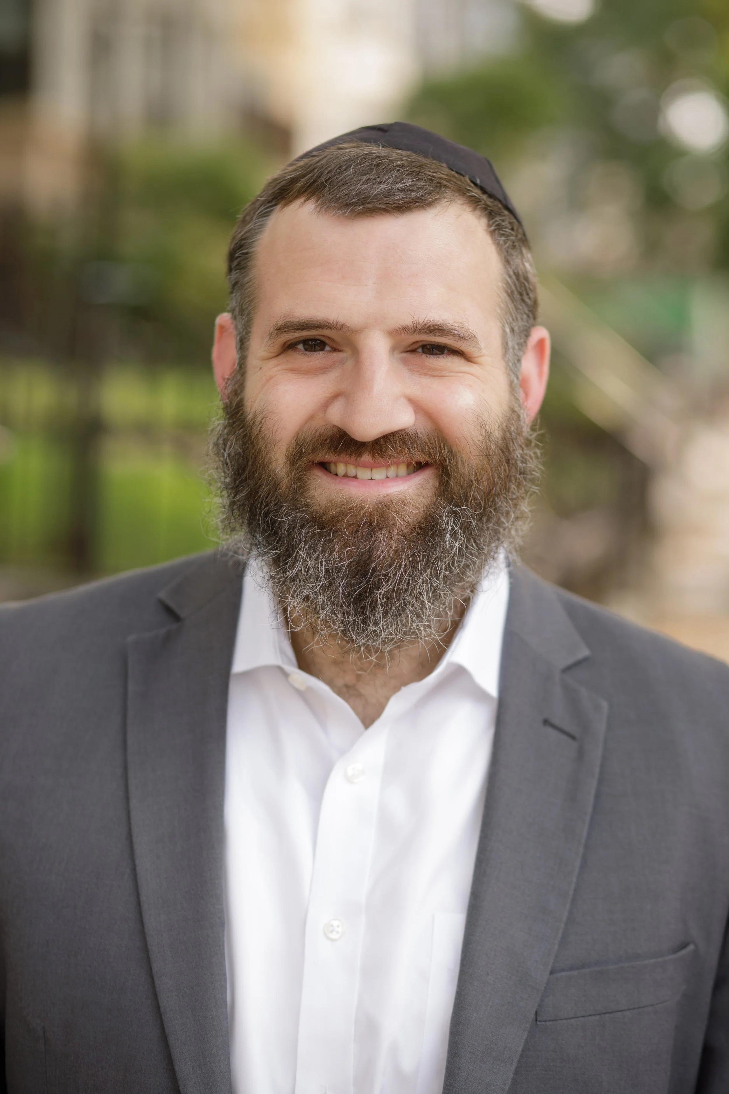 A man with a beard wearing a black kippah, a white dress shirt, and a gray suit jacket, smiling outdoors with a blurred background of trees and buildings.