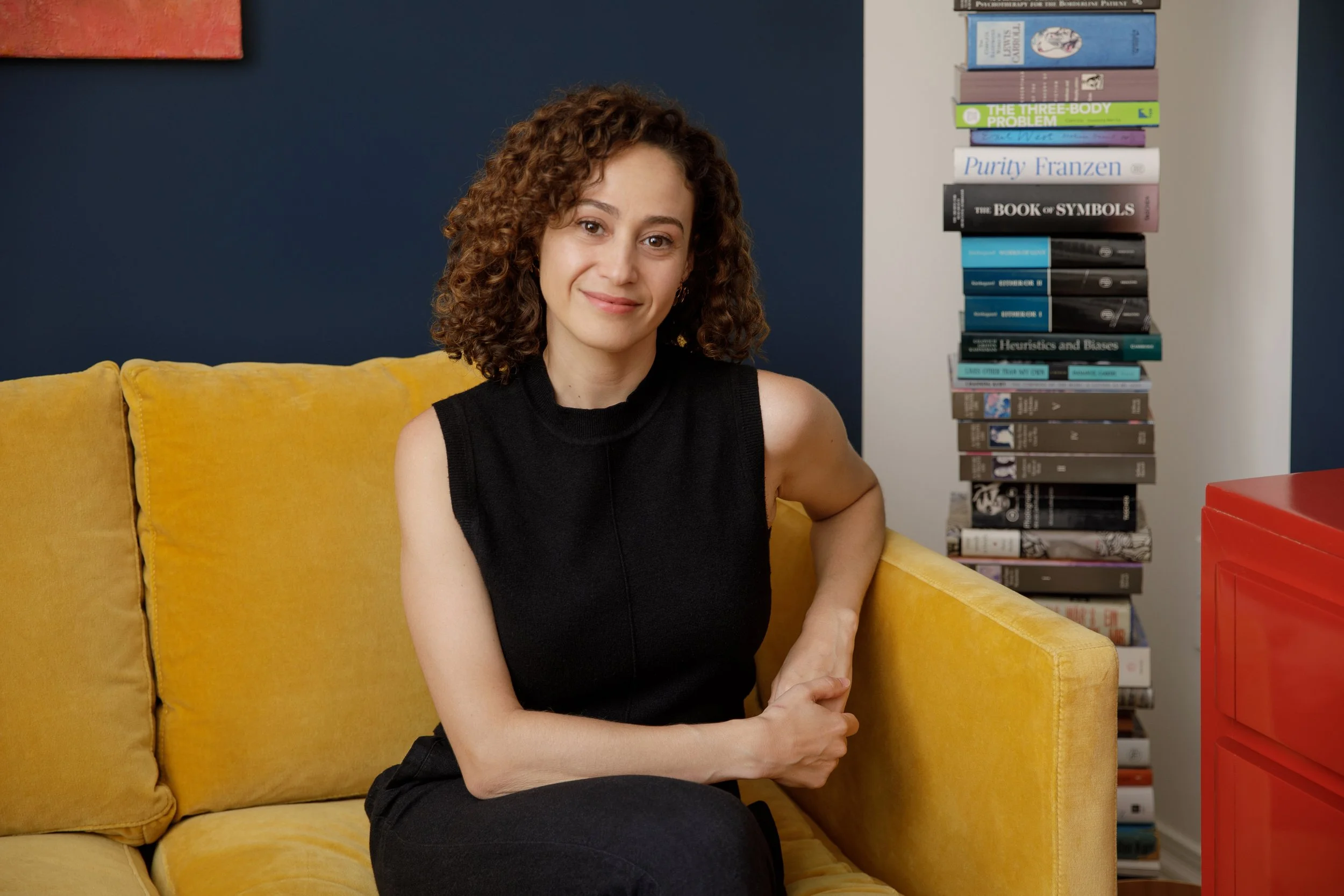A woman with curly hair dressed in black, sitting on a yellow couch, smiling softly at the camera in a room with a bookshelf and a red box.