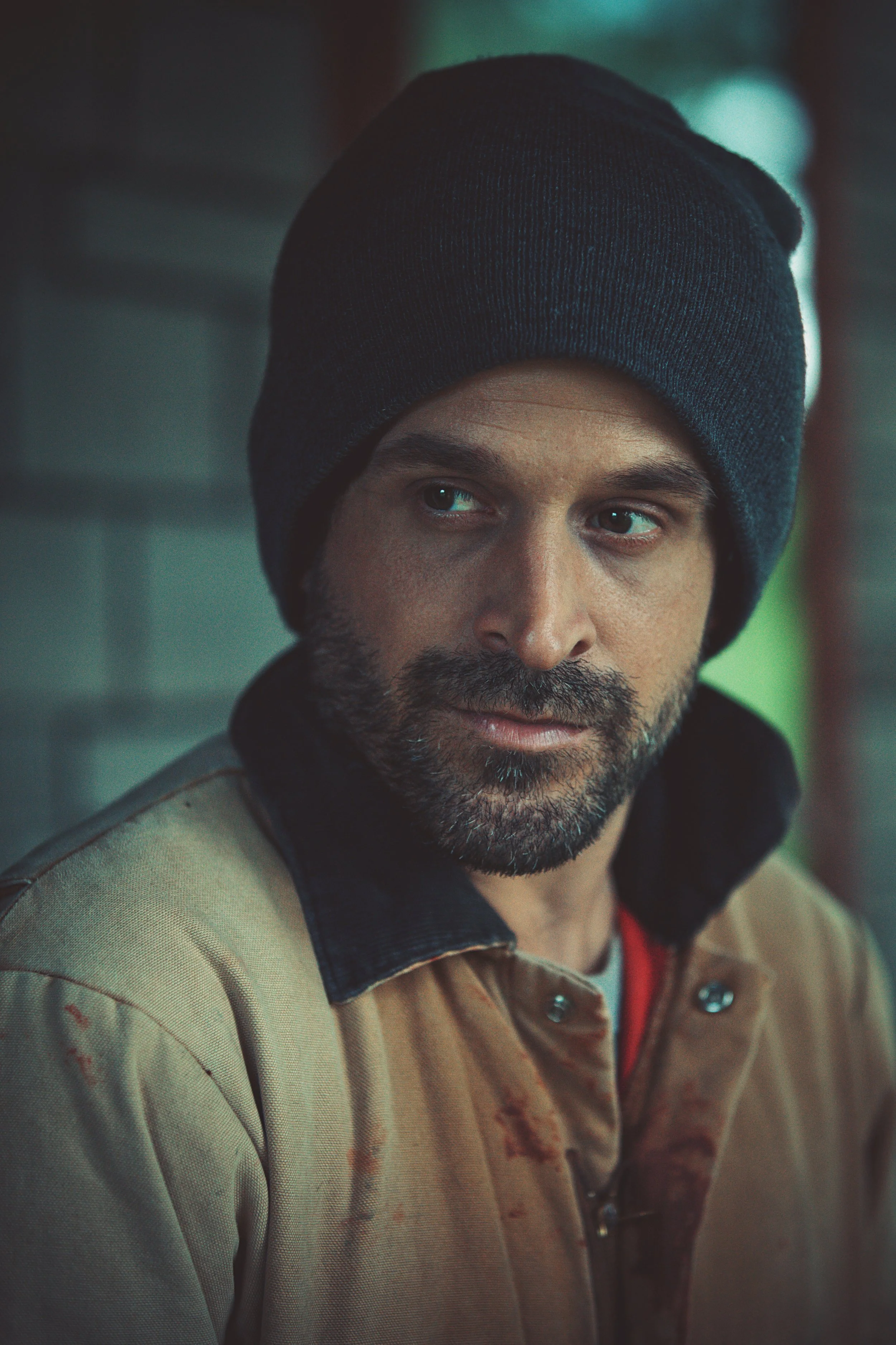 Close-up of a man with dark hair, a beard, wearing a black beanie, a tan jacket, and a hoodie inside. He is looking to the side with a serious expression.