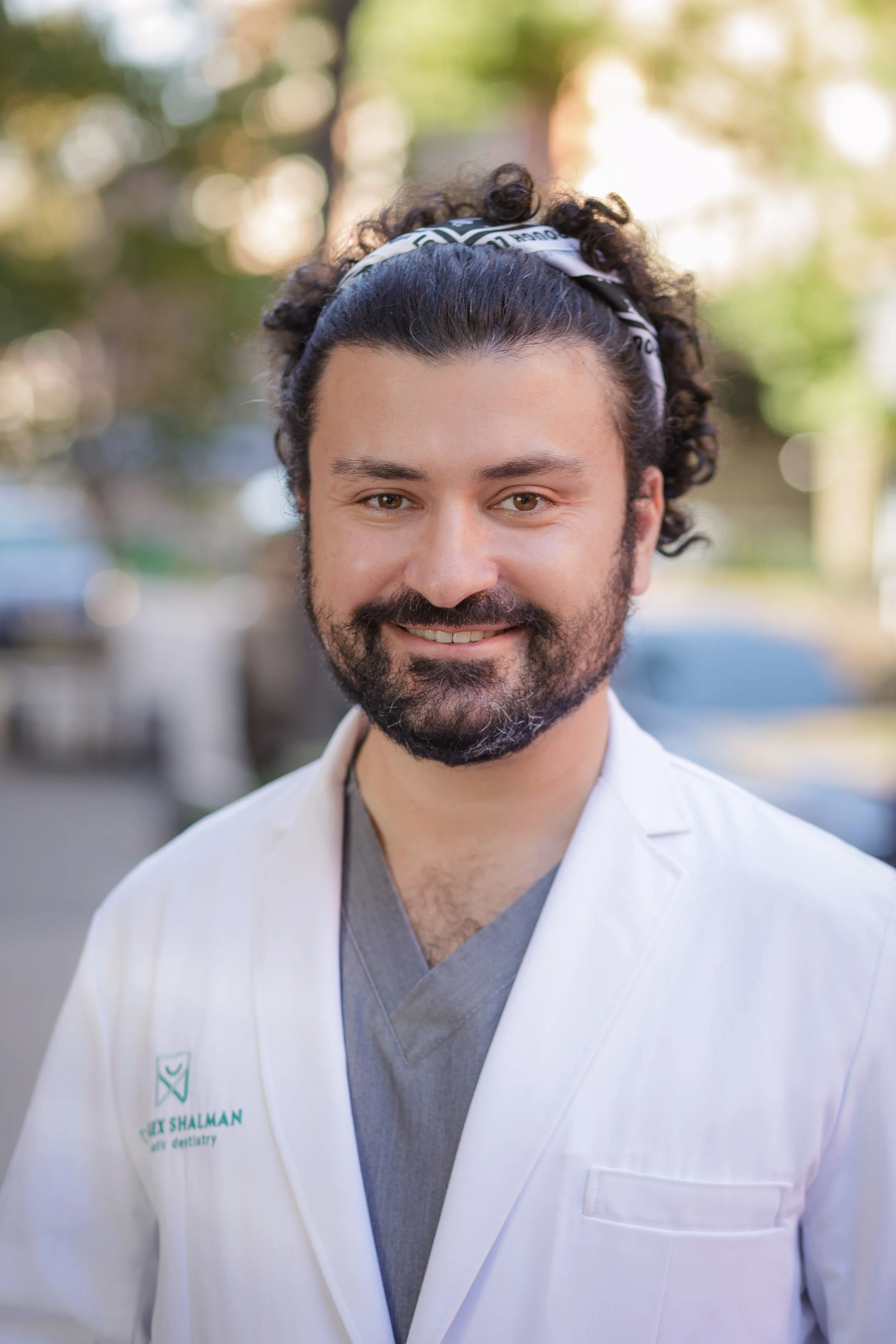 A man with dark, curly hair and a beard, wearing a white medical coat and gray scrub top, standing outdoors, smiling, with blurred trees and sunlight in the background.