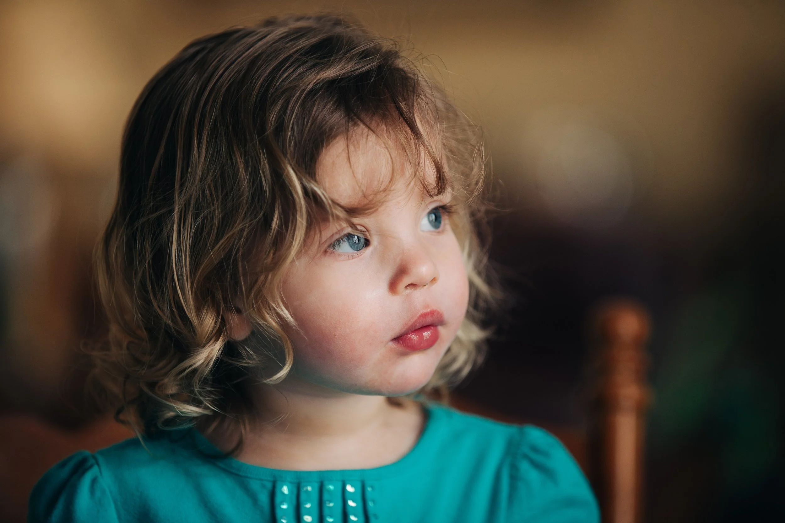 Close-up of a young girl with curly blonde hair and blue eyes, wearing a teal shirt, looking to the side with a thoughtful expression.