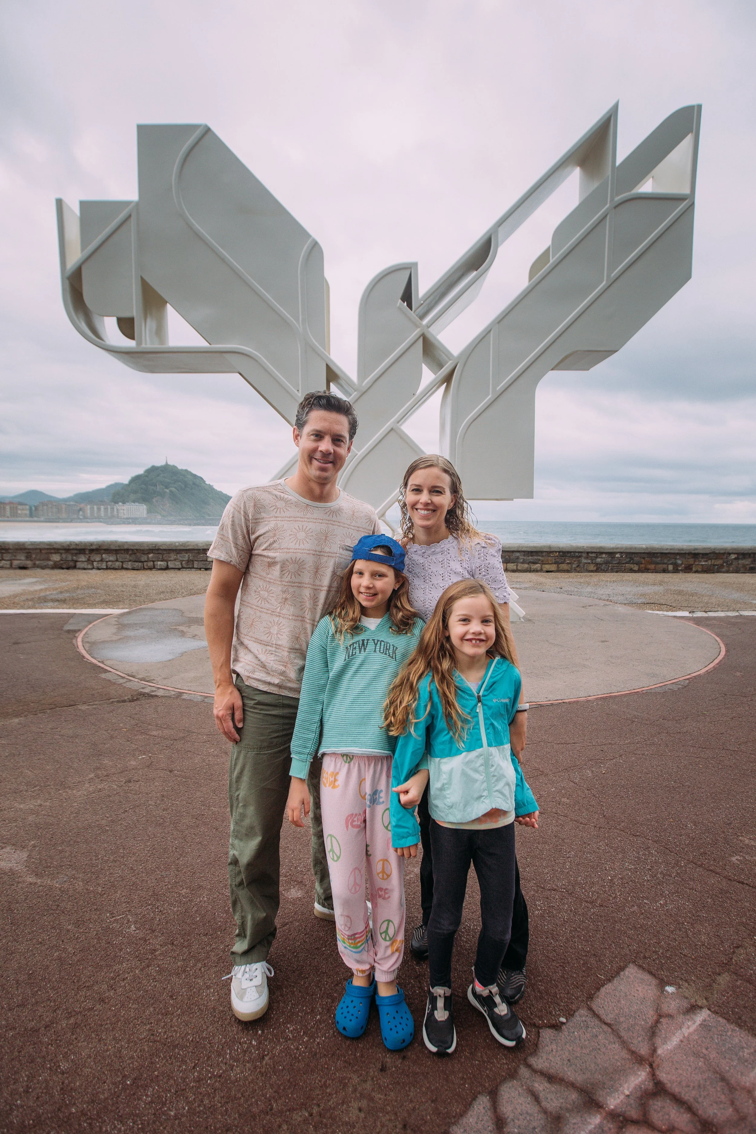 A family of five standing outdoors near the ocean with a large white abstract sculpture behind them. The family includes a man, a woman, and three young girls. The sky is cloudy and there is a hilly landscape with buildings in the background.
