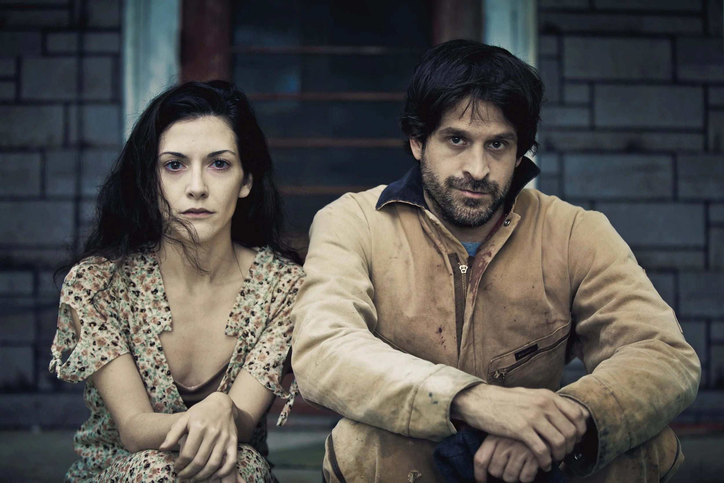 A man and woman sitting outdoors on wooden stairs, with brick wall background. The woman has long black hair, wearing a floral dress, and has a serious expression. The man has dark hair and beard, wearing a beige jacket, and also has a serious expres