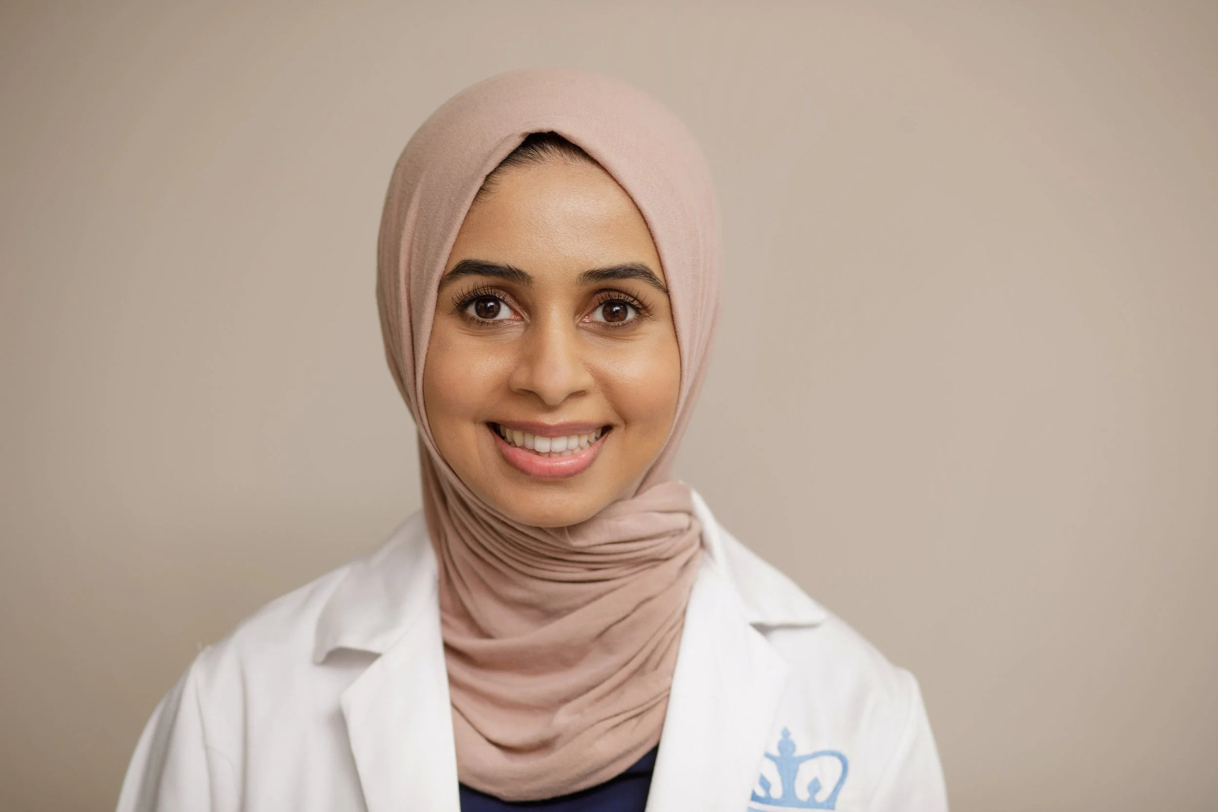 Portrait of a smiling woman wearing a pink hijab and white lab coat against a plain beige background.