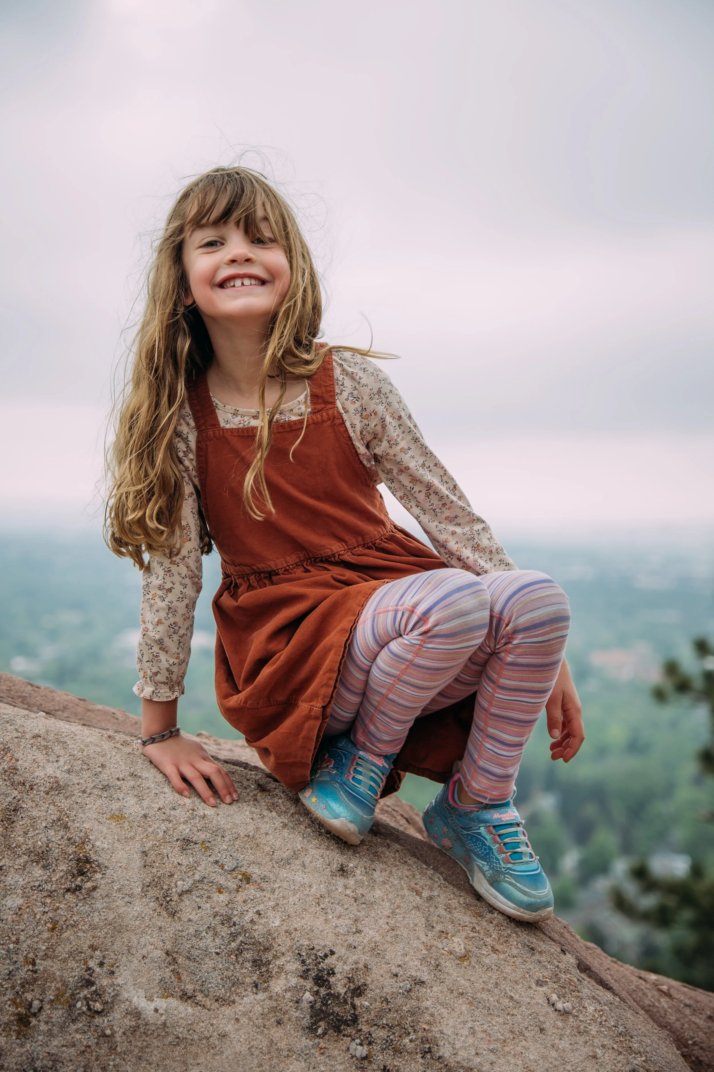 A young girl with long, wavy brown hair, smiling and sitting on a rocky surface outdoors, wearing a brown dress with a floral long-sleeve shirt underneath, striped leggings, and blue sneakers, with a scenic blurred landscape in the background.