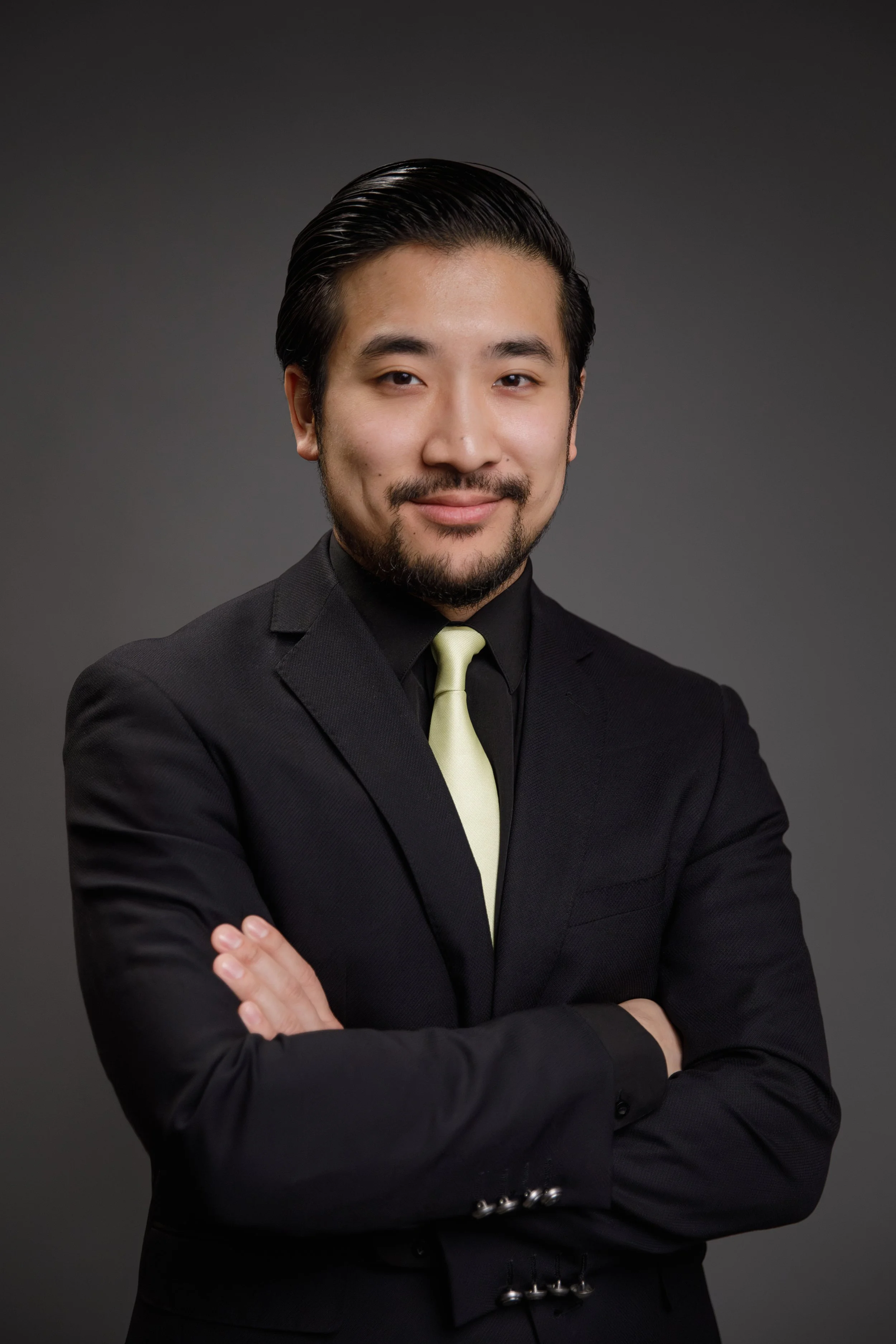 Professional portrait of a man with dark hair and a trimmed beard, wearing a black suit, black shirt, and a light-colored tie, standing with arms crossed against a dark background.