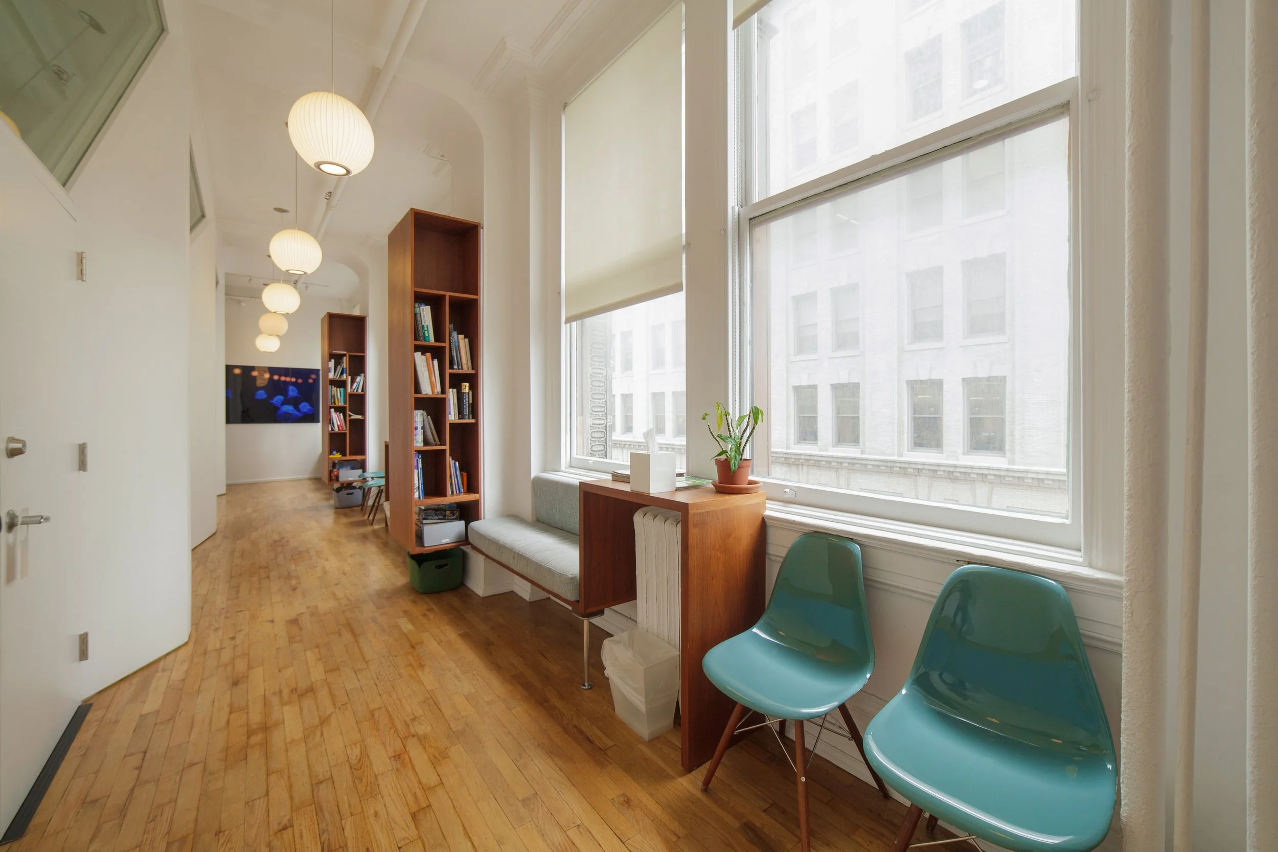 A bright room with large windows and white walls. There are wooden bookshelves filled with books, a small table with a potted plant, and blue chairs near the windows. Overhead, spherical pendant lights hang from the ceiling.