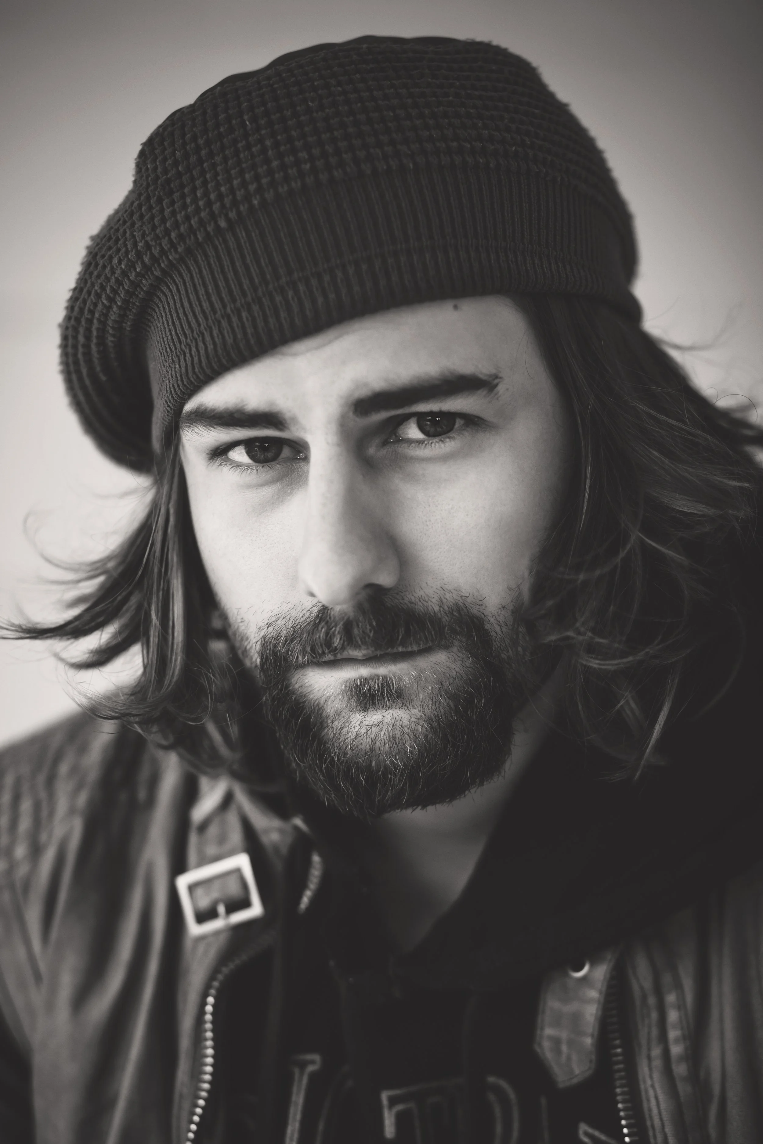 Black and white close-up portrait of a young man with long hair, beard, and mustache wearing a knit beanie and a leather jacket.