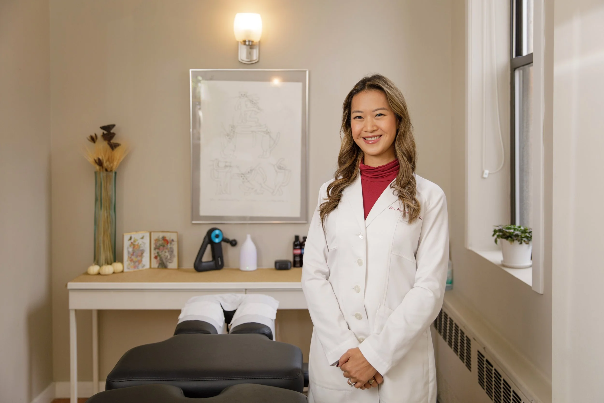A female healthcare professional in a white coat standing in a medical office with a treatment table, decorative artwork, and a window in the background.