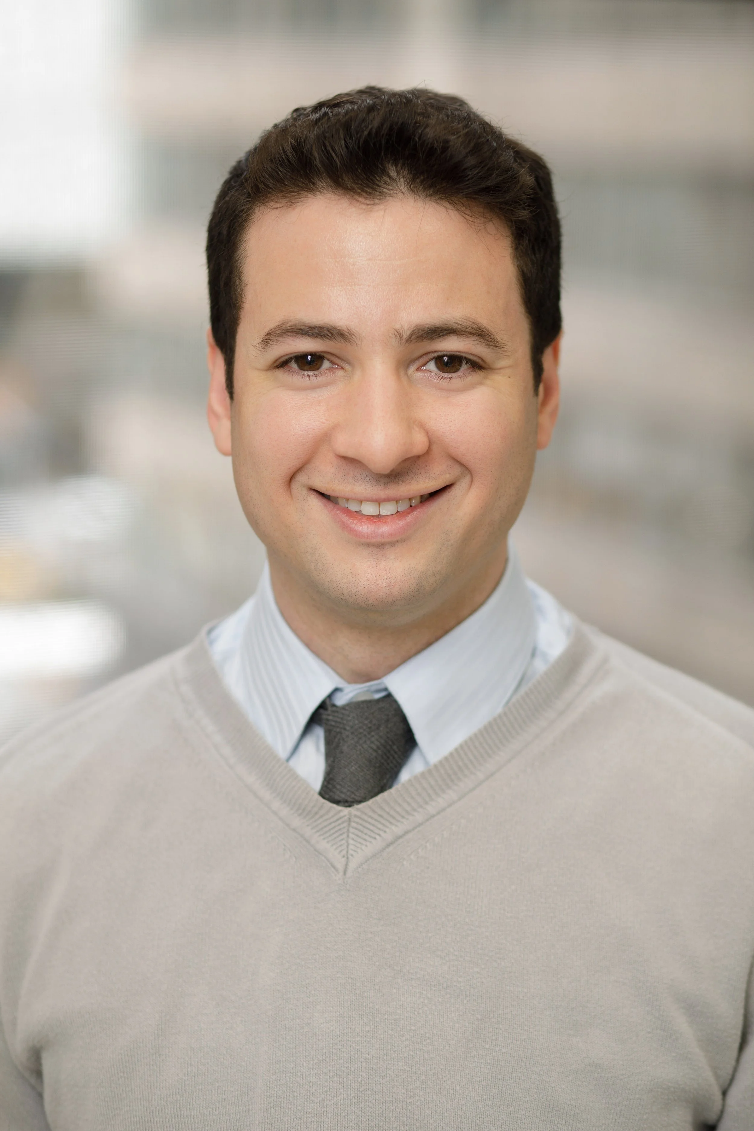 Headshot of a smiling man with dark hair in business casual attire, standing indoors with a blurred background.