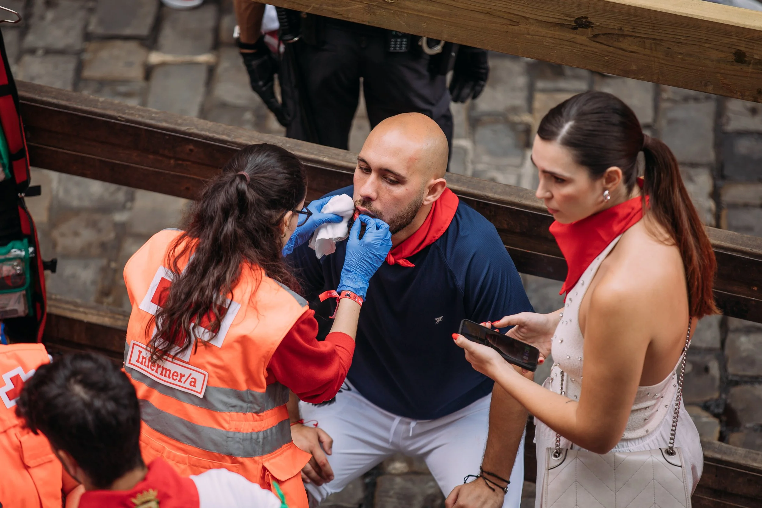 An emergency medical worker administering aid to a man with a shaved head and beard, with two women standing nearby, one holding a smartphone, in an outdoor setting with stone steps.