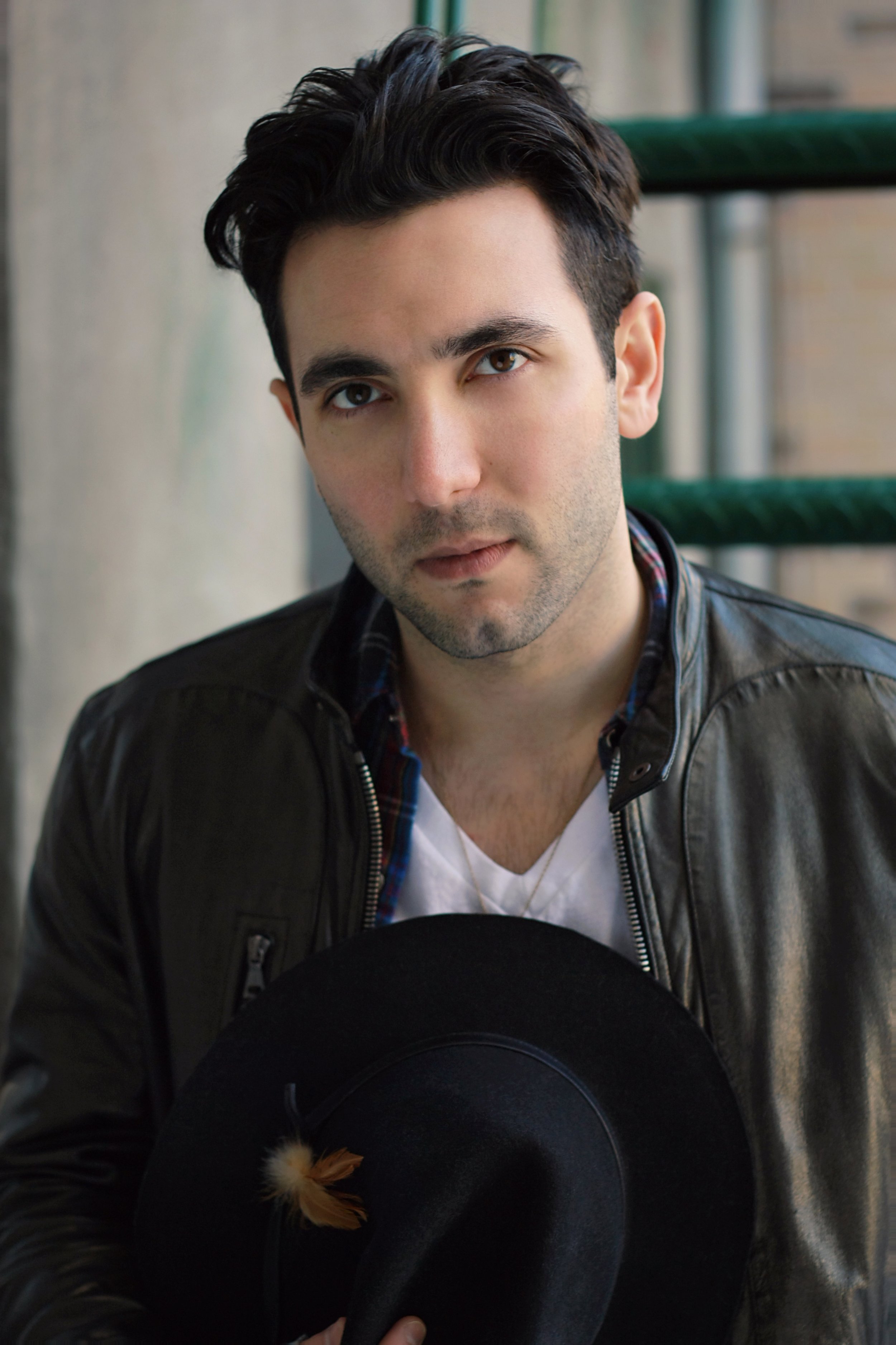 A young man with dark hair and brown eyes, wearing a black leather jacket and a white T-shirt, standing outside or near a building, with a green railing and a blurred background.