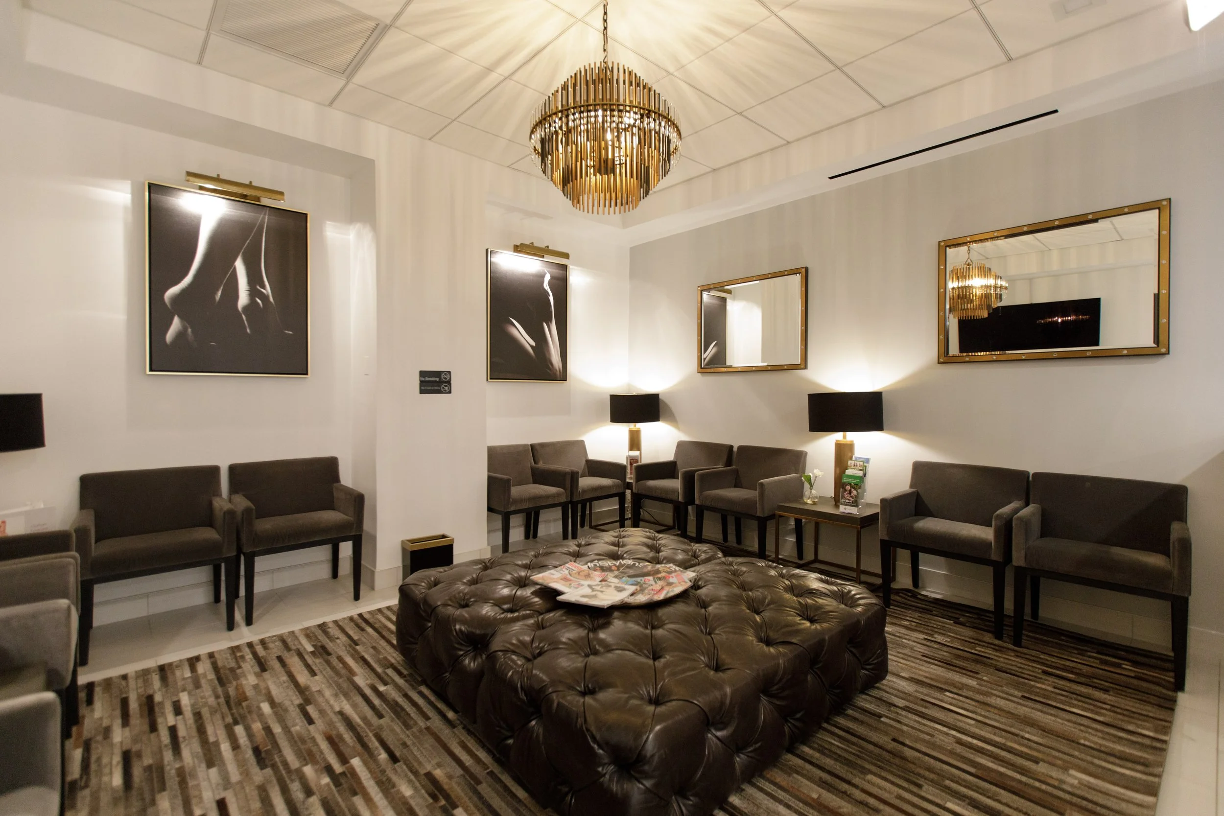A modern lounge area with multiple grey armchairs arranged along the walls, a large brown leather tufted ottoman in the center with magazines on top, and a striped area rug on the floor. The white walls are decorated with black and white framed artwo