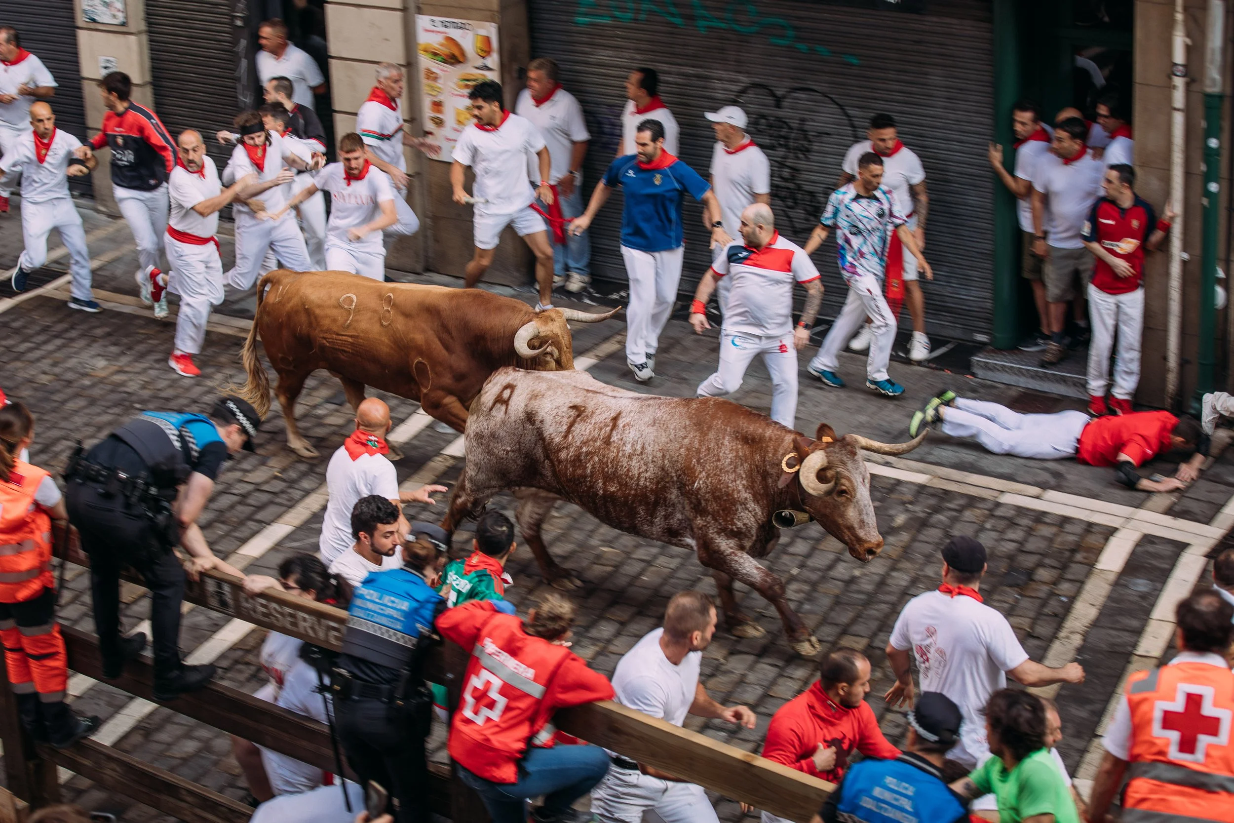 People participating in a running of the bulls event in an urban street; some people are running while others are lying on the ground or being restrained for safety around bulls.